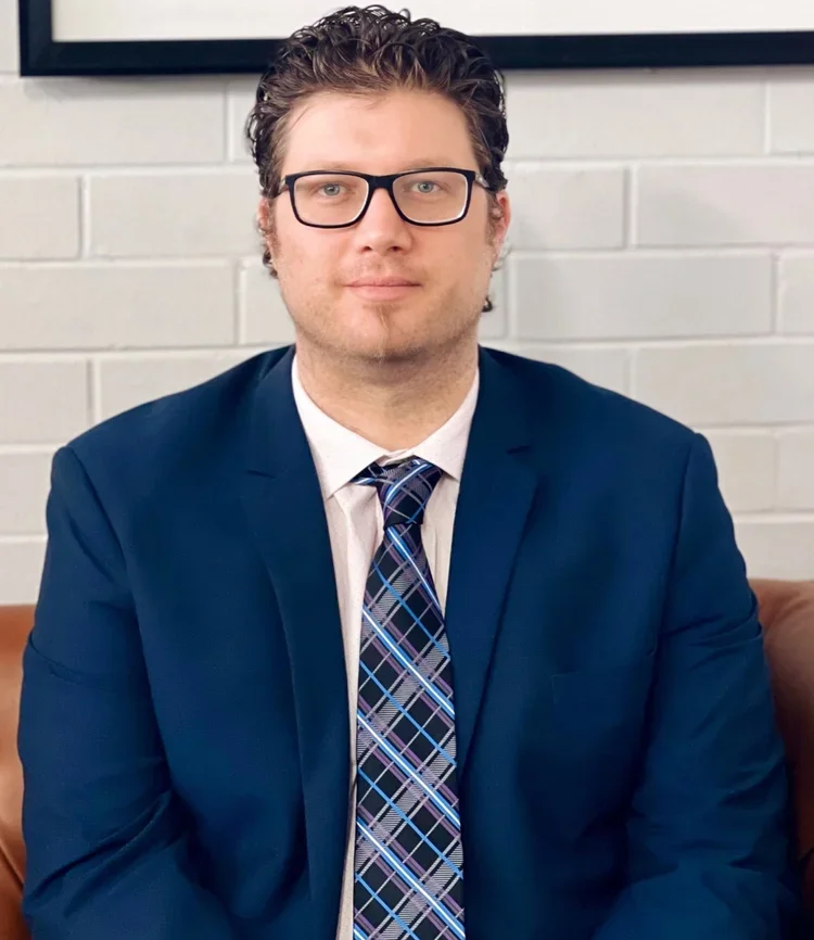 A man in a dark blue suit, white shirt, and a blue plaid tie, wearing glasses, sitting against a white brick wall.