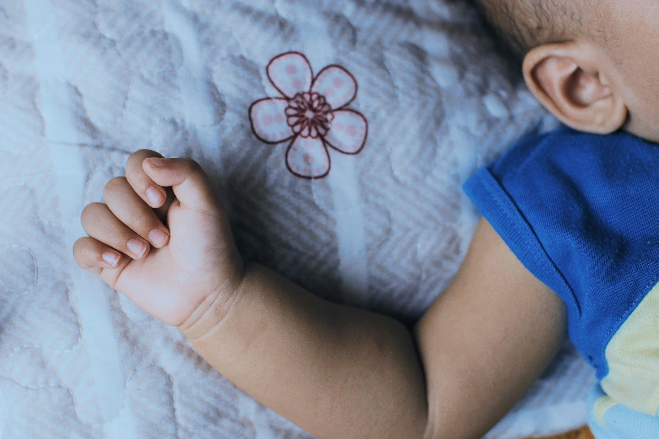 Close-up of a child's hand resting on a patterned surface with a flower design, part of the child's face and shoulder visible, wearing a blue and yellow shirt.
