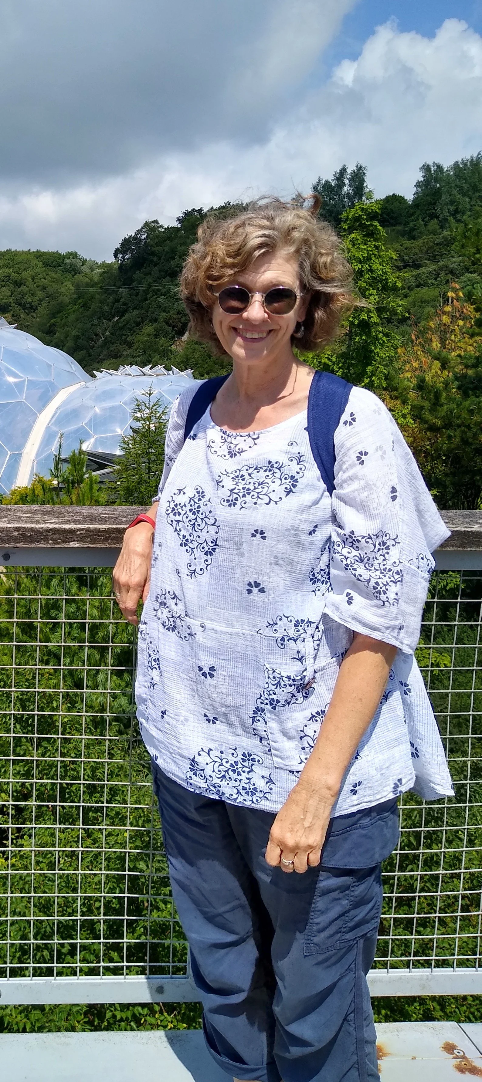 Smiling woman standing outdoors on a bridge with green trees and a cloudy sky in the background.