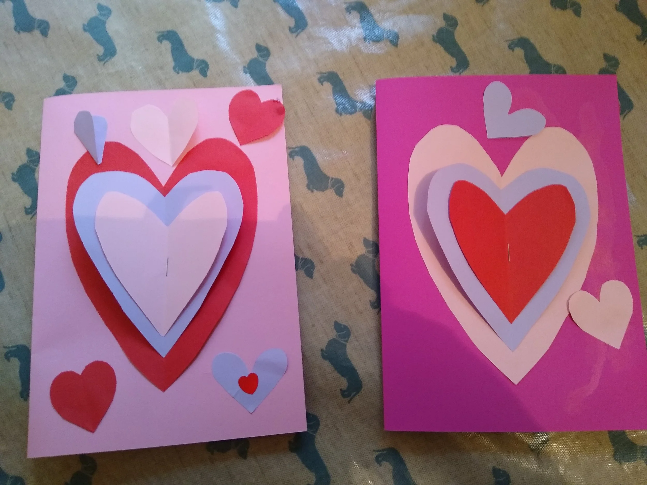 Two handmade Valentine's Day cards decorated with paper hearts, one pink and one purple, each featuring layered paper hearts and additional smaller hearts.