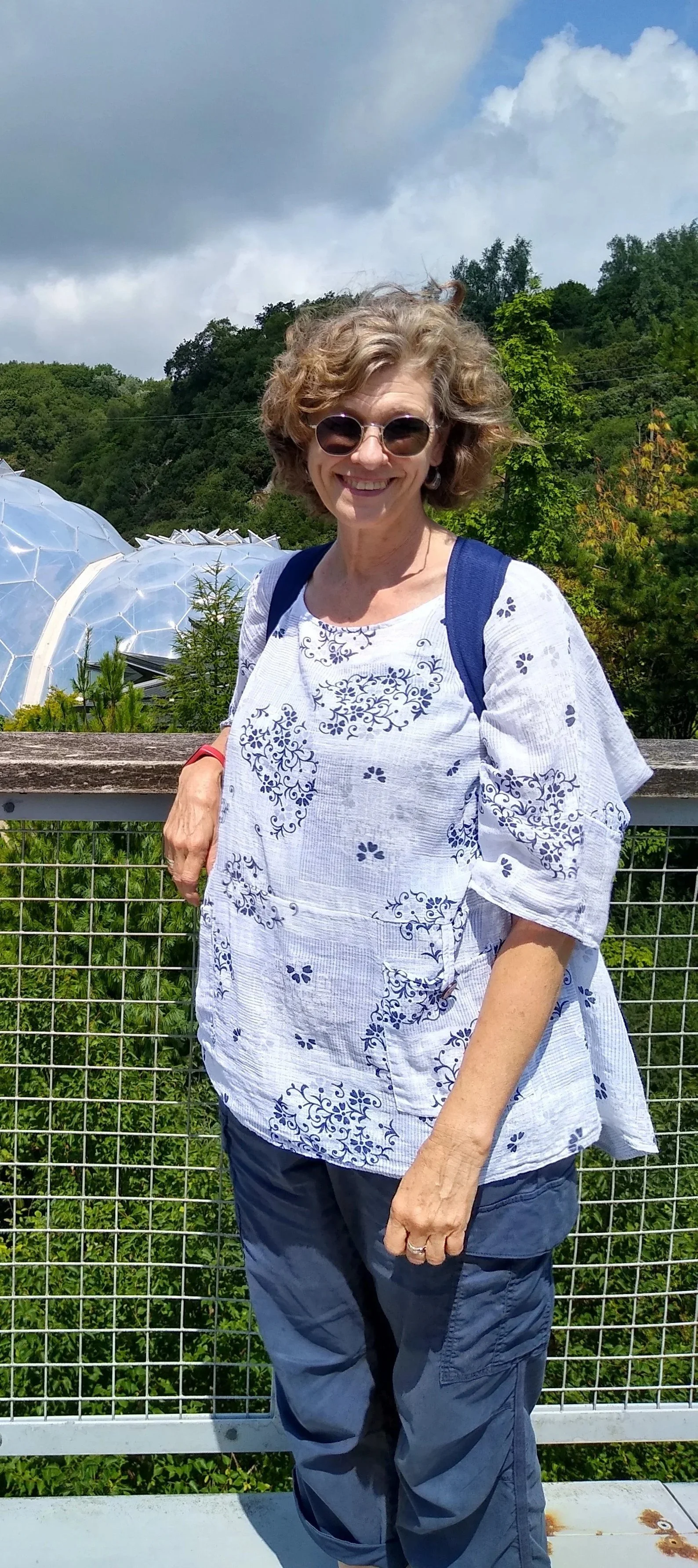 A woman with curly blonde hair and sunglasses smiling outdoors on a cloudy day, wearing a white blouse with blue embroidery and blue pants, standing in front of greenery and a greenhouse.