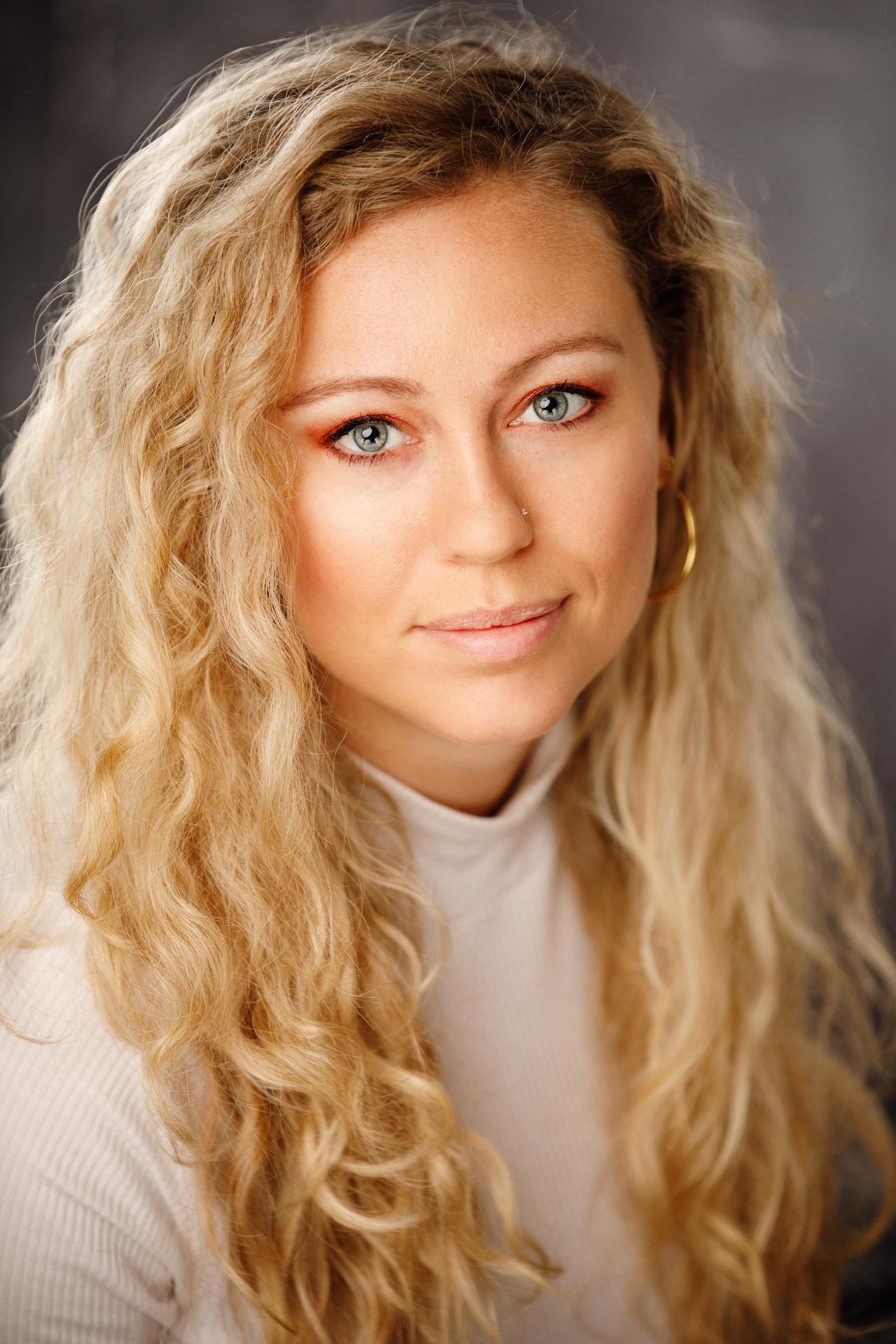 A fresh, close-crop studio portrait of a blonde woman with natural curly hair and blue eyes, wearing a white turtleneck against a soft grey backdrop. Gold hoop earrings, minimal makeup, warm and open expression. No heavy styling, no dramatic contrast