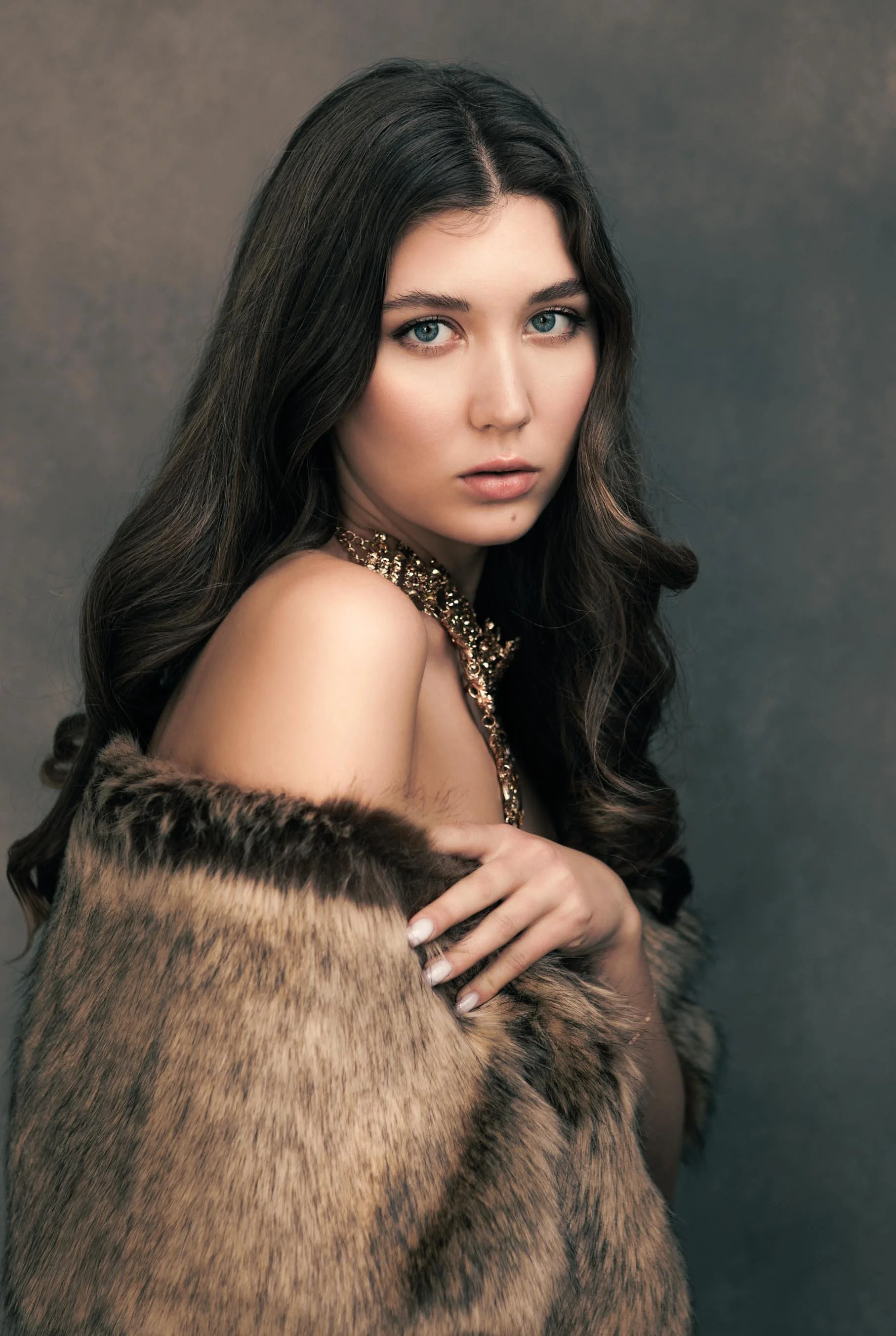 A fine art studio portrait of a young woman with long dark hair, draped in a fur stole with a gold statement necklace against a textured grey backdrop. Off-shoulder composition, direct gaze, bare skin against rich texture. The image sits firmly in lu