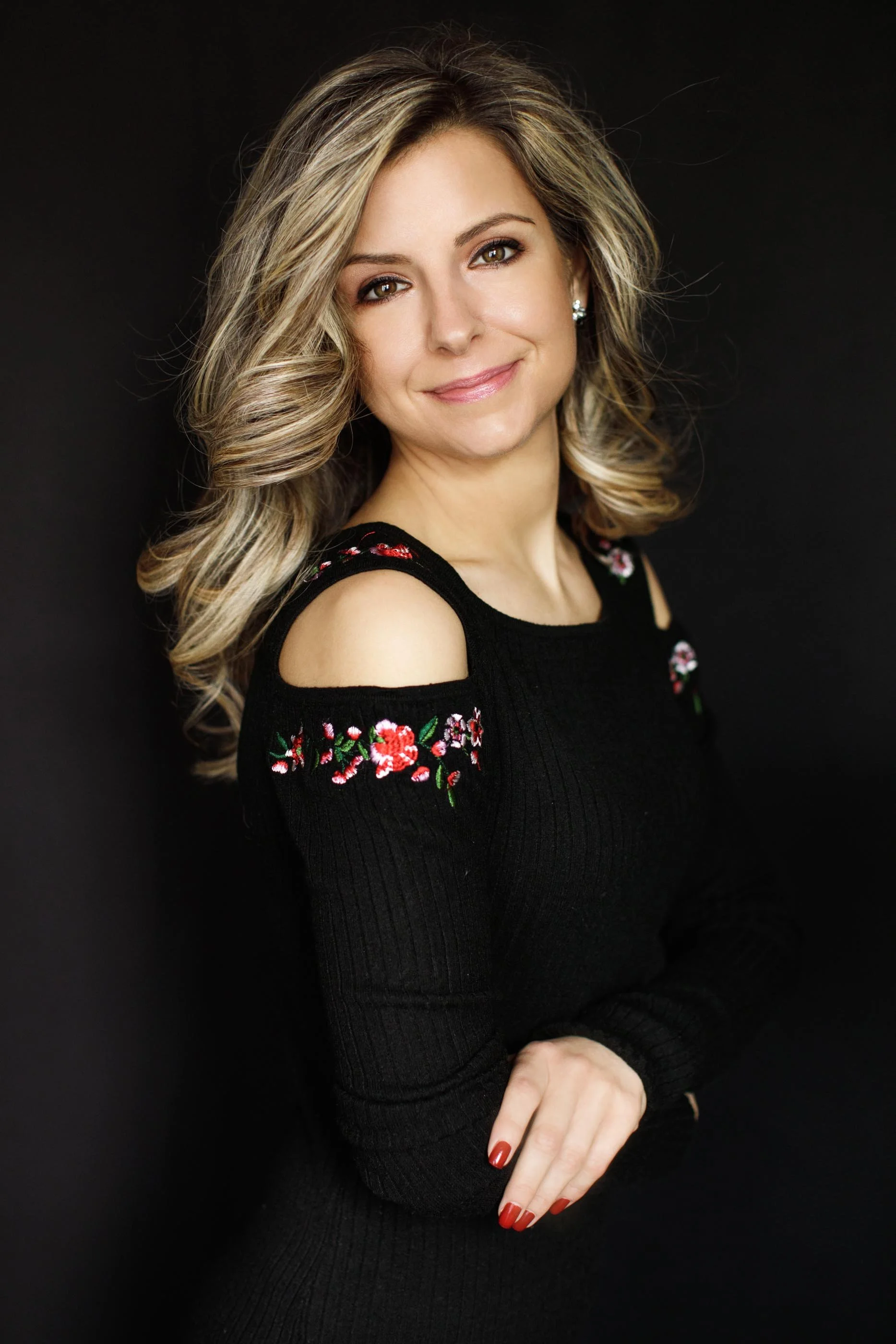 A woman with blonde, wavy hair wearing a black, long-sleeved sweater with floral embroidery on the shoulders and arms, standing against a dark background, smiling at the camera.