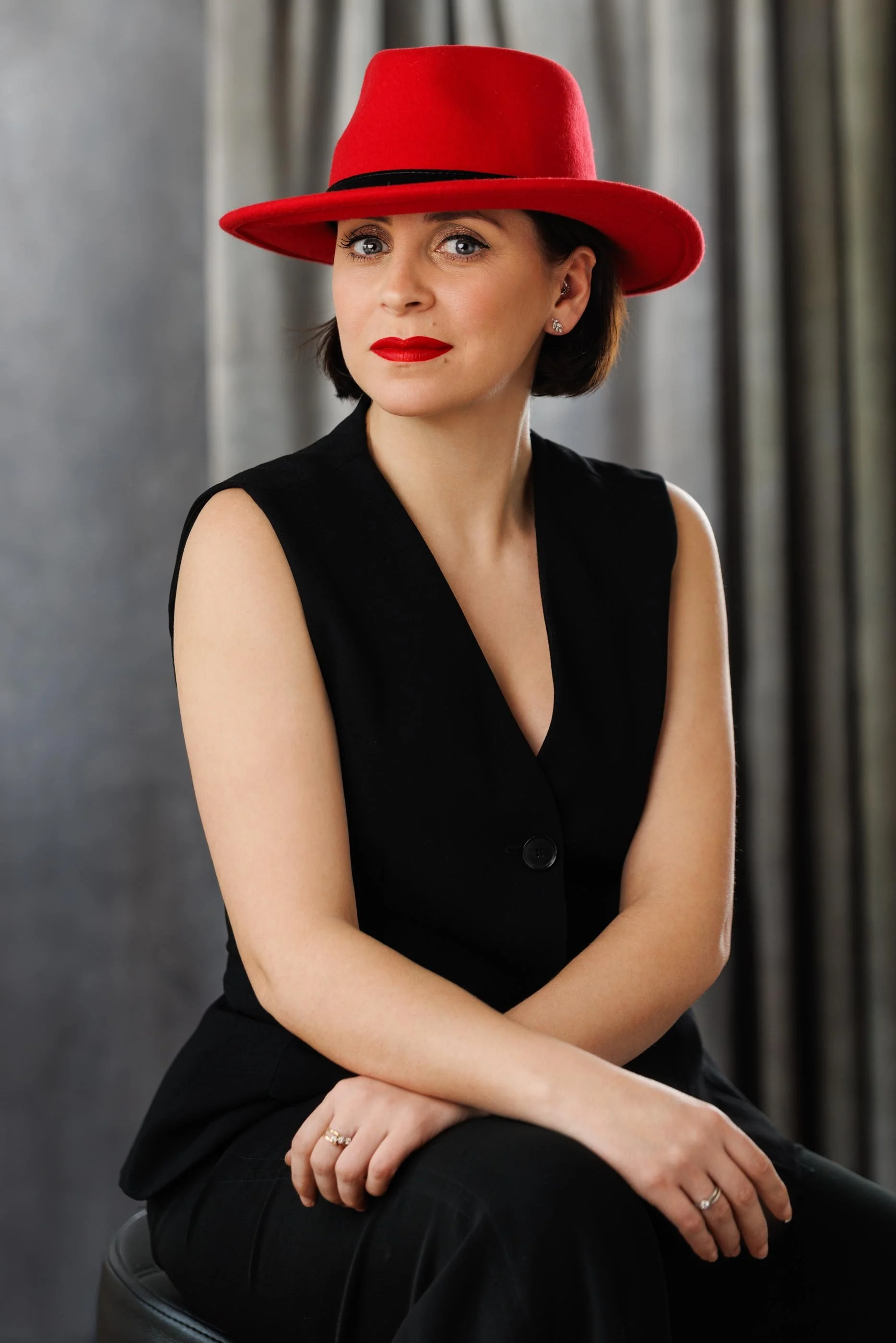 A strong studio portrait of a woman in a black sleeveless suit and bold red fedora, seated against a grey draped backdrop. Red lip, direct gaze, hands relaxed in lap. Controlled and deliberate — every element of the frame is working. This is the kind