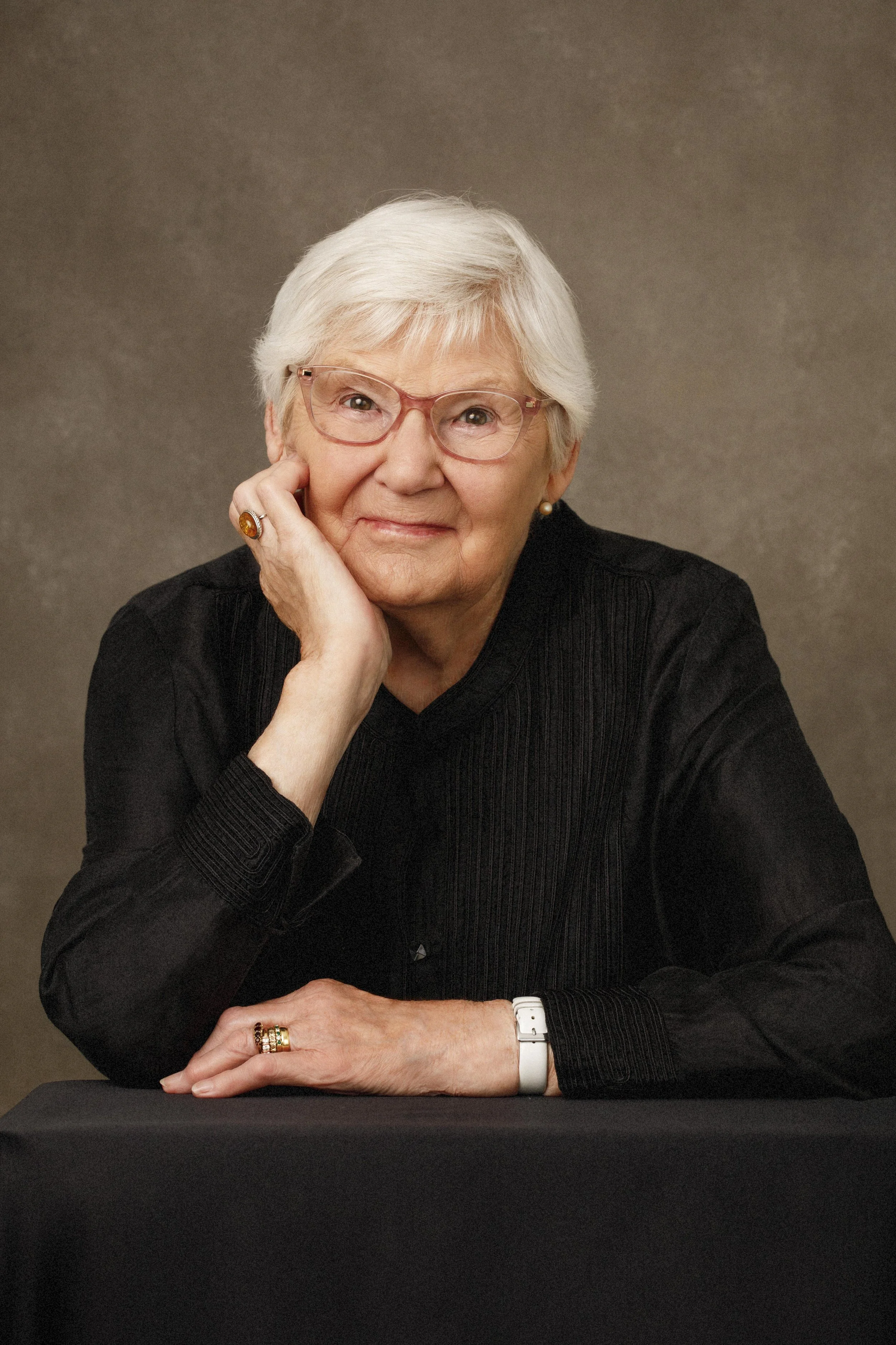 Elegant older woman wearing glasses and a black dress with hand wrapping her face and looking into camera directly photographed in  a private studio in East Lothian by Angelique Portrait Photography