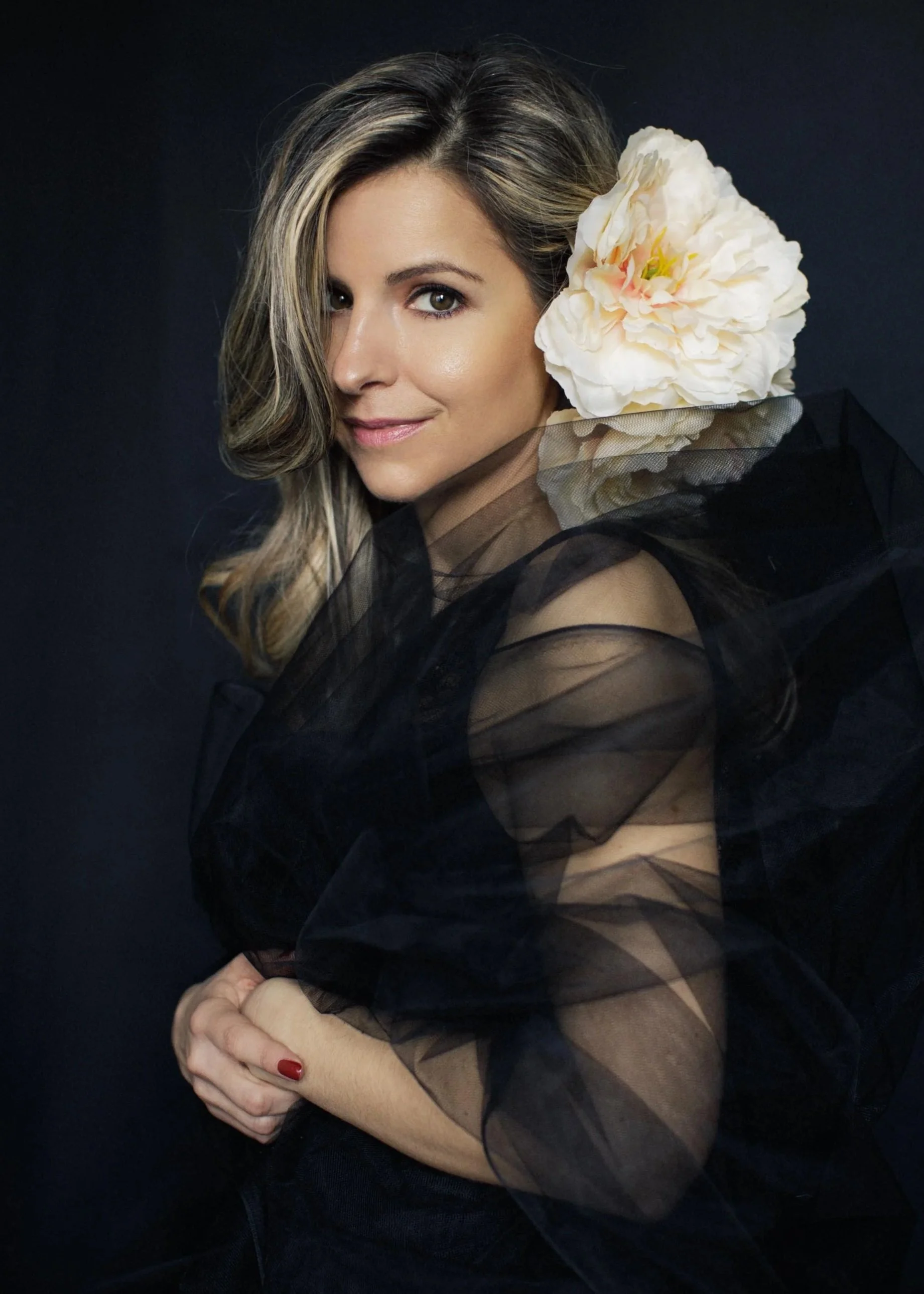 A dramatic, fine art studio portrait of a silver-blonde woman wrapped in sheer black fabric with a large white peony at her shoulder, shot against a deep dark backdrop. Arms crossed, slight smile, assured presence. This image and the darker floral po