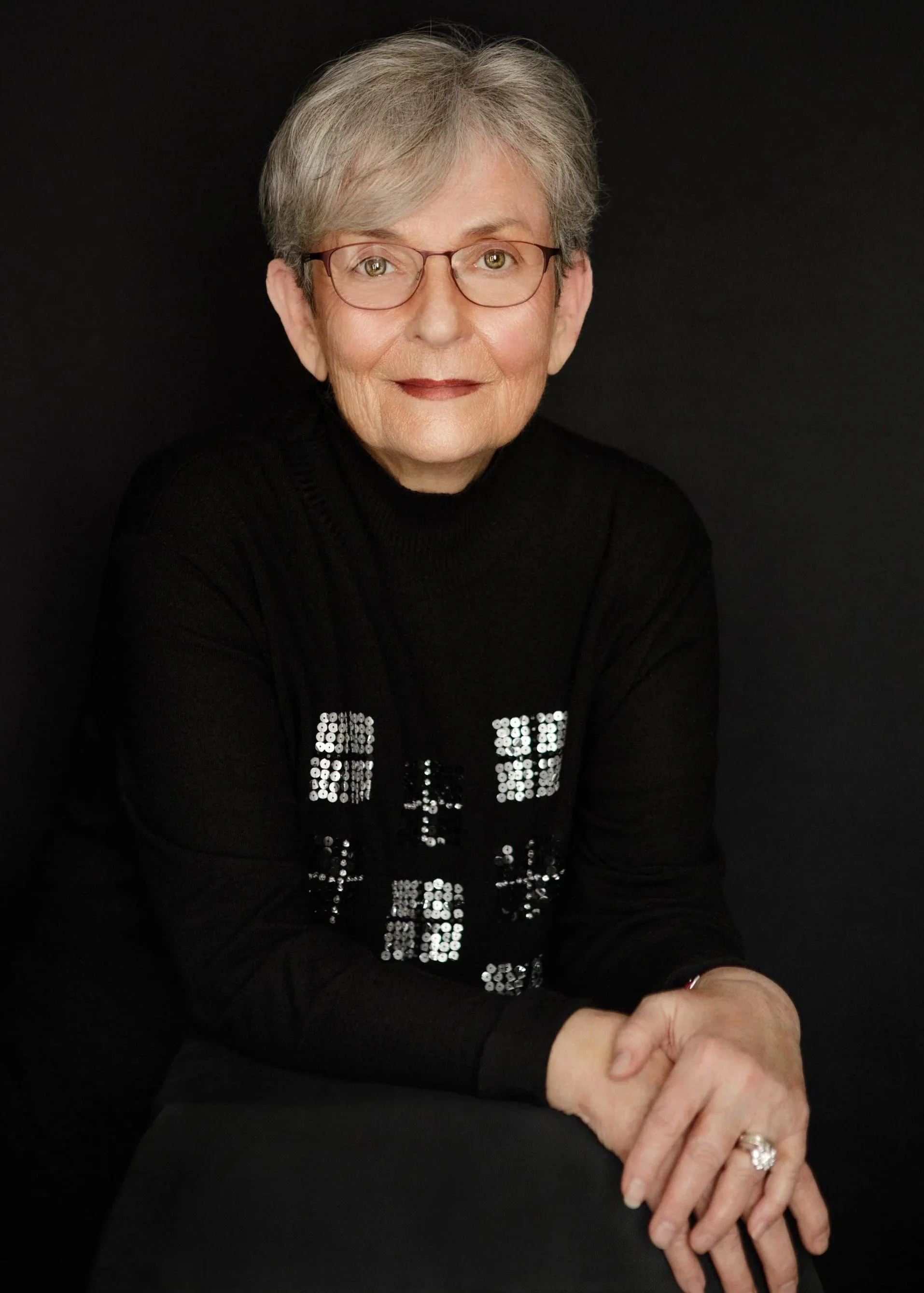 A refined studio portrait of a woman with cropped silver hair and glasses, wearing a black turtleneck with sequin detail. Hands folded, direct gaze, dark backdrop. Understated elegance. This image speaks directly to women who know exactly who they ar