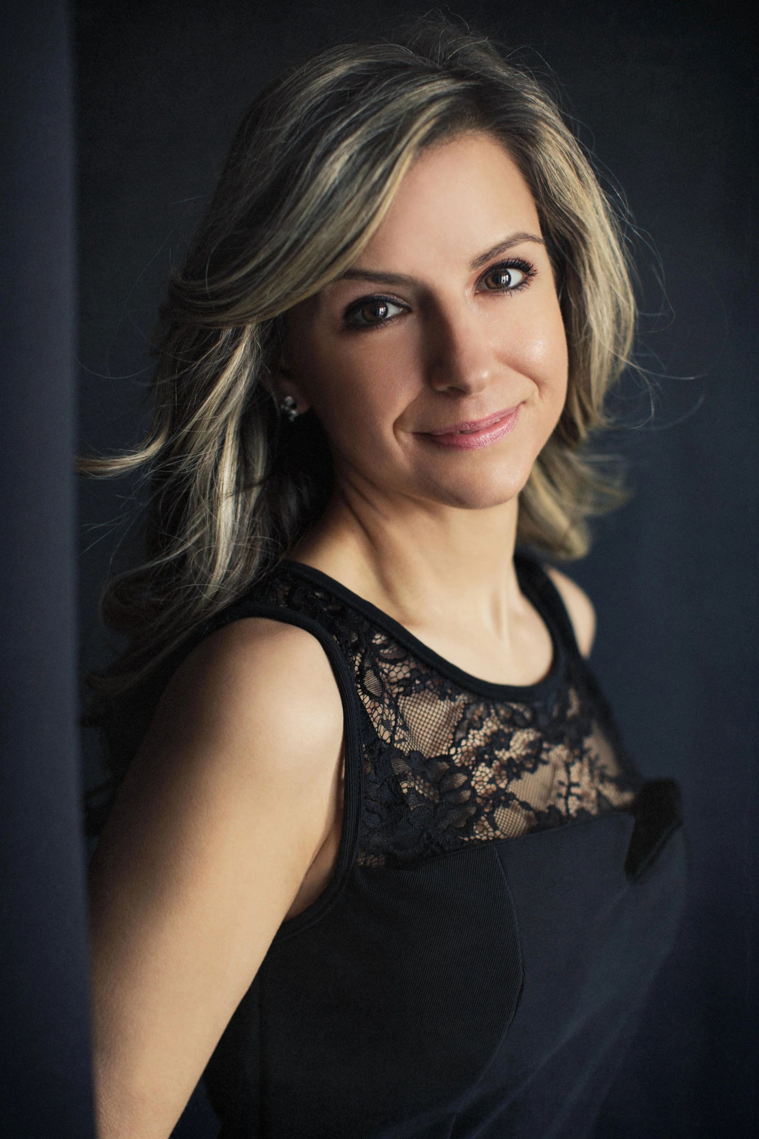 A classic studio portrait of a woman with silver-blonde hair, wearing a black lace dress. Soft directional lighting, dark backdrop. Warm, composed expression — approachable authority. The kind of image that works equally on a corporate biography page