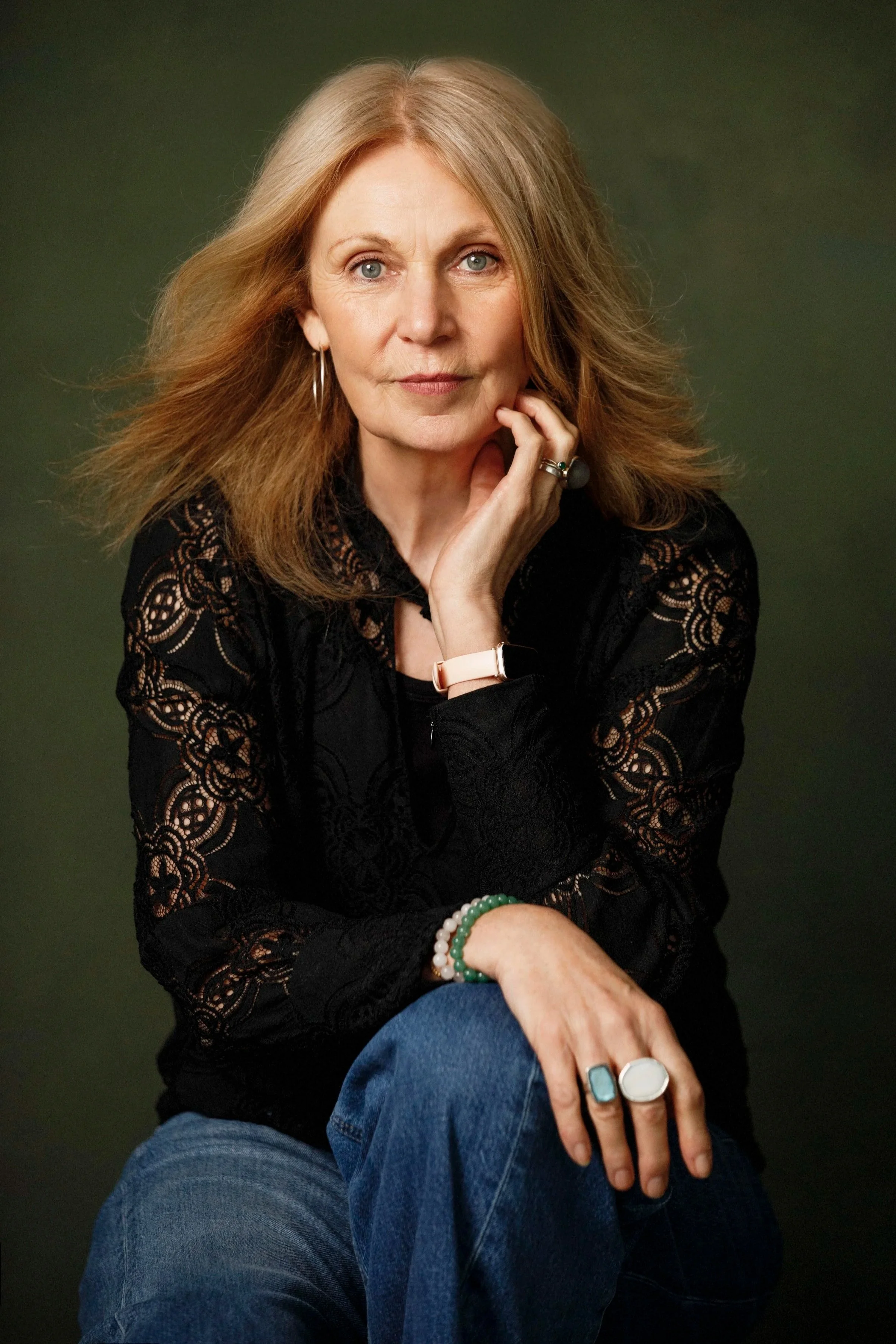 A woman with long blonde hair, blue eyes, and light skin is sitting against a dark green background. She is wearing a black lace long-sleeve top, blue jeans, and various rings and bracelets. She is resting her chin on her hand, looking directly at th