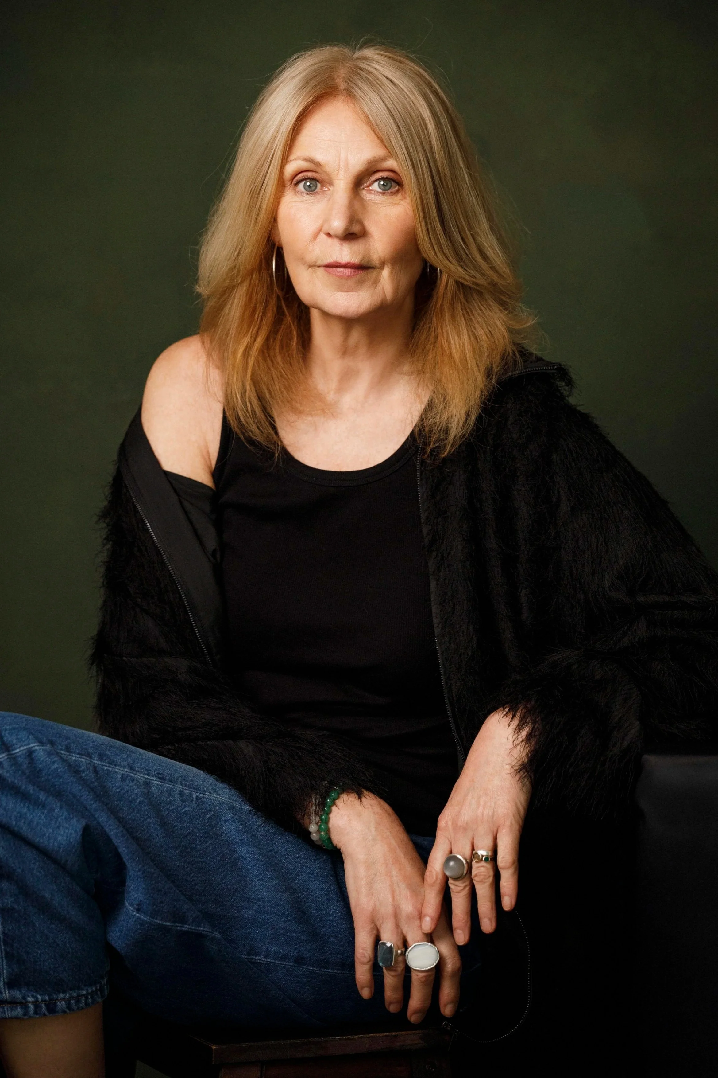 Close-up portrait of a middle-aged woman with blonde hair and blue eyes, wearing a black top and rings, sitting against a grayish-green background.