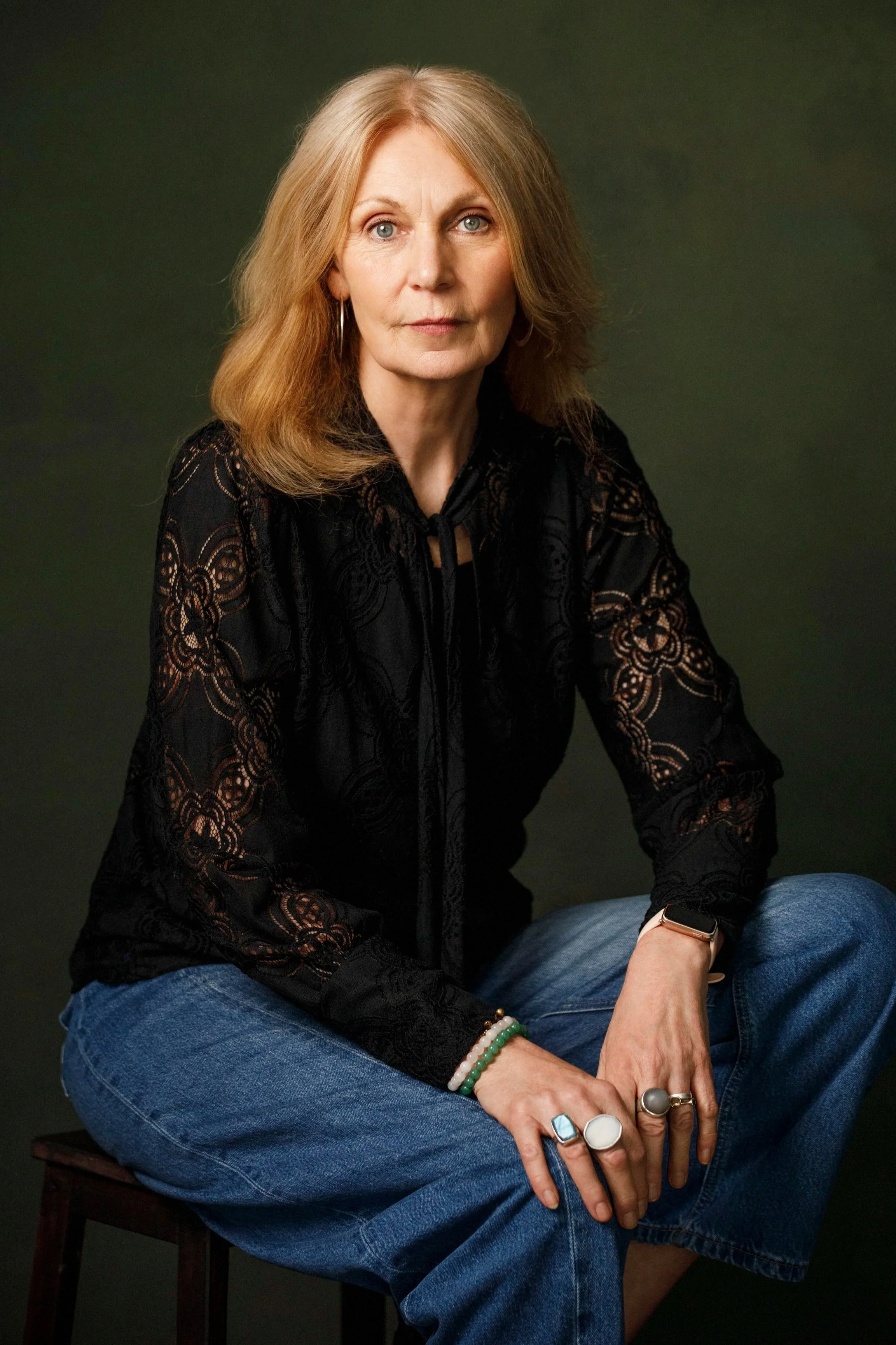 A woman with long reddish-brown hair and light skin sitting on a wooden chair against a dark green background. She is wearing a black lace blouse, blue jeans, and multiple rings and bracelets, with a smartwatch on her left wrist. She has blue eyes an