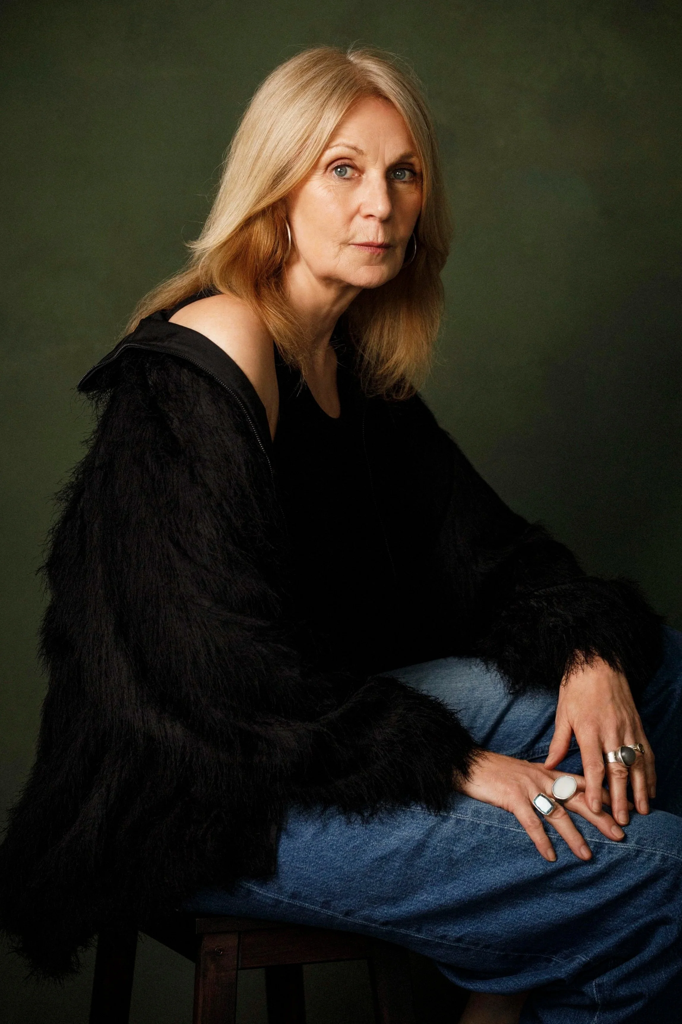 A woman with long light brown hair and blue eyes, wearing a black fuzzy knit cardigan and multiple silver rings on her fingers, sitting on a wooden chair against a dark background.