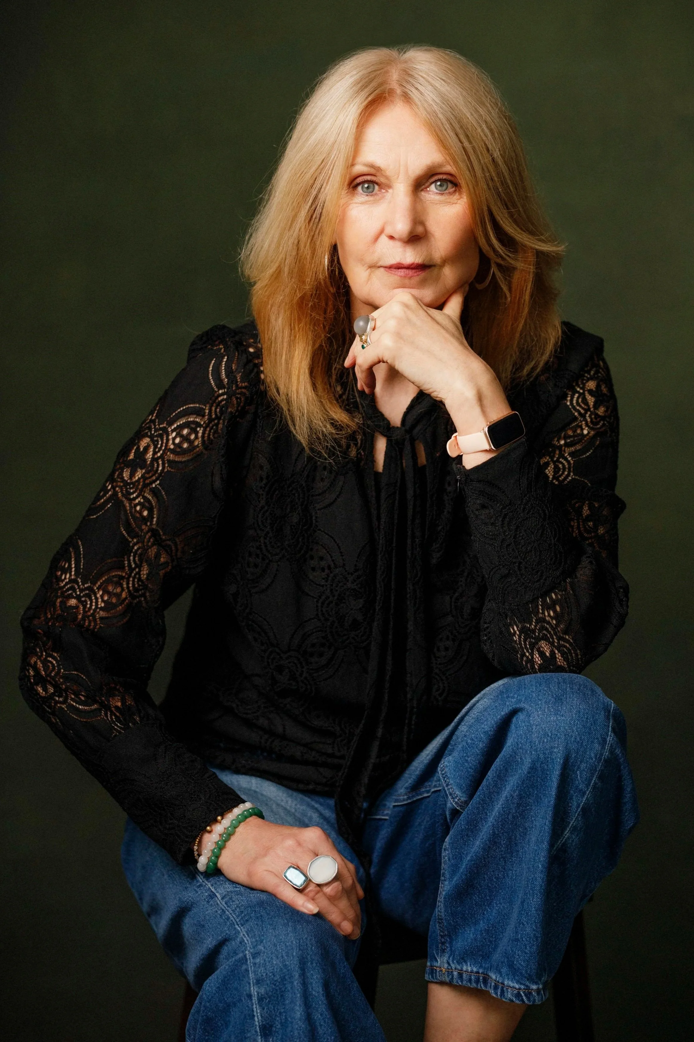 A woman with blonde hair and blue eyes wearing a black lace top and blue jeans, sitting with one leg crossed over the other, resting her chin on her hand, wearing various rings and bracelets, and an Apple Watch on her left wrist, against a dark green