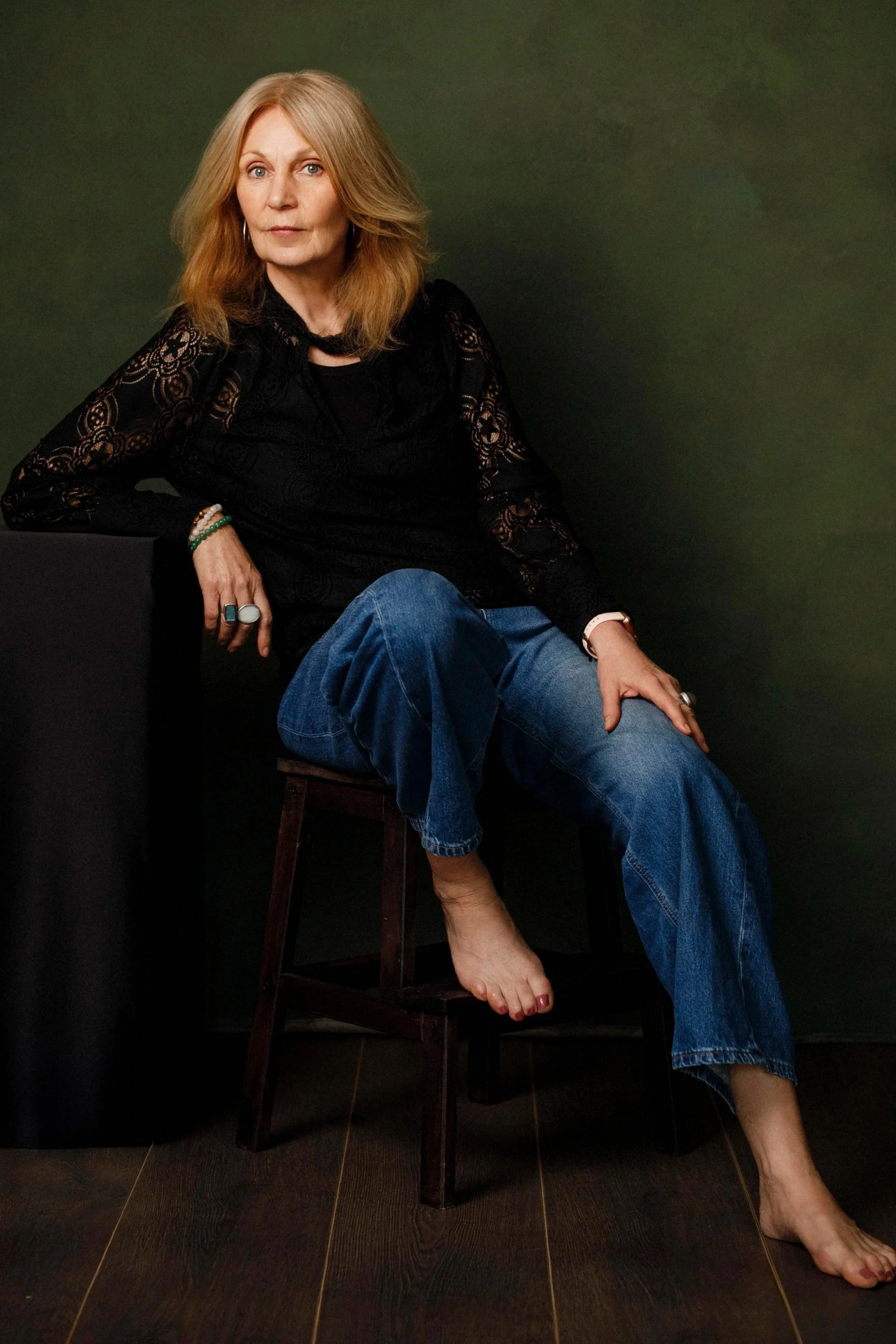 A woman with long red hair sitting on a wooden stool against a green background, wearing a black lace top and blue jeans, with her right arm resting on a black piece of furniture, accessorized with rings and bracelets, and barefoot.