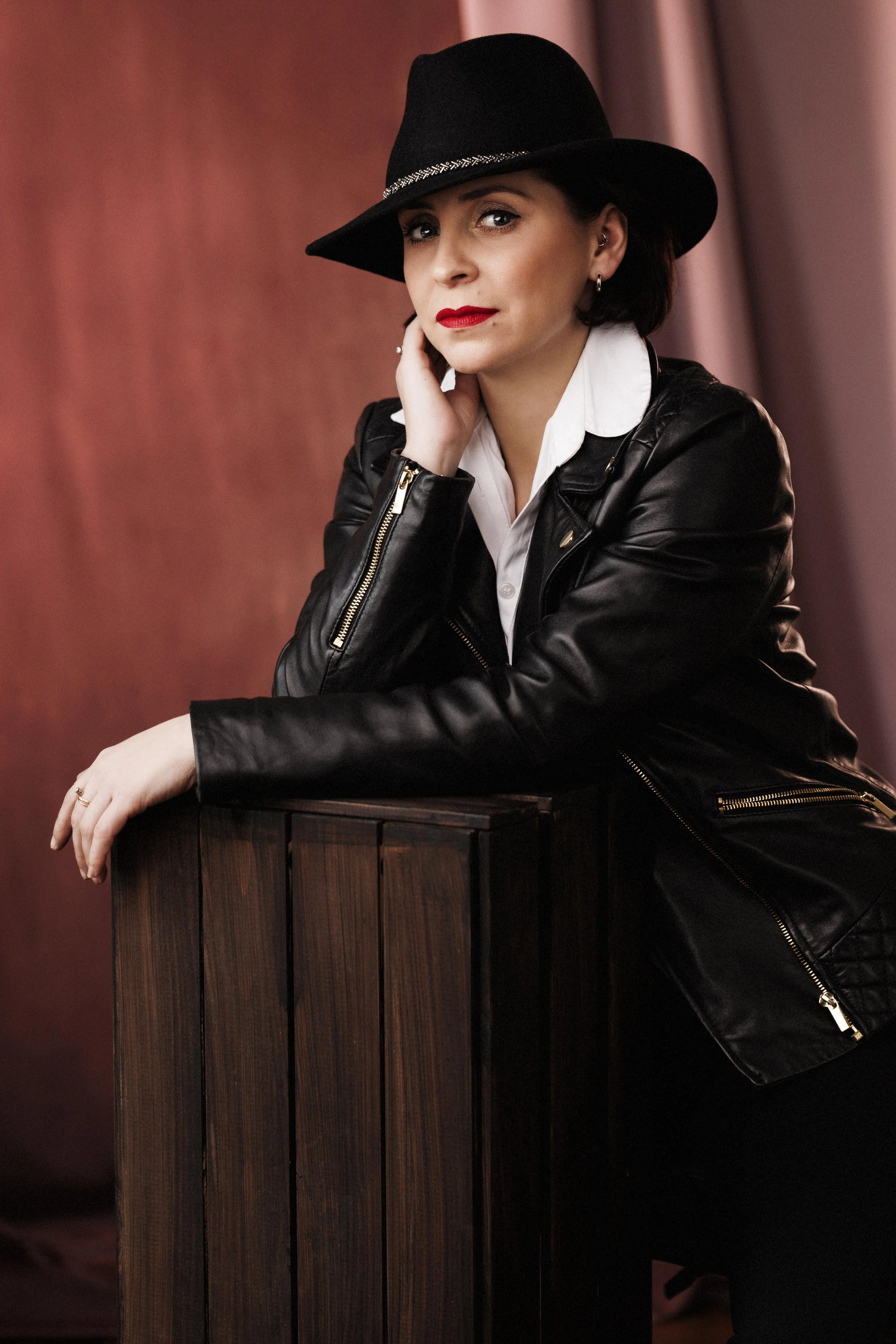 Elegant woman wearing black fedora hat wearing leather black jacket and gorgeous red lipstick, looking directly into camera.