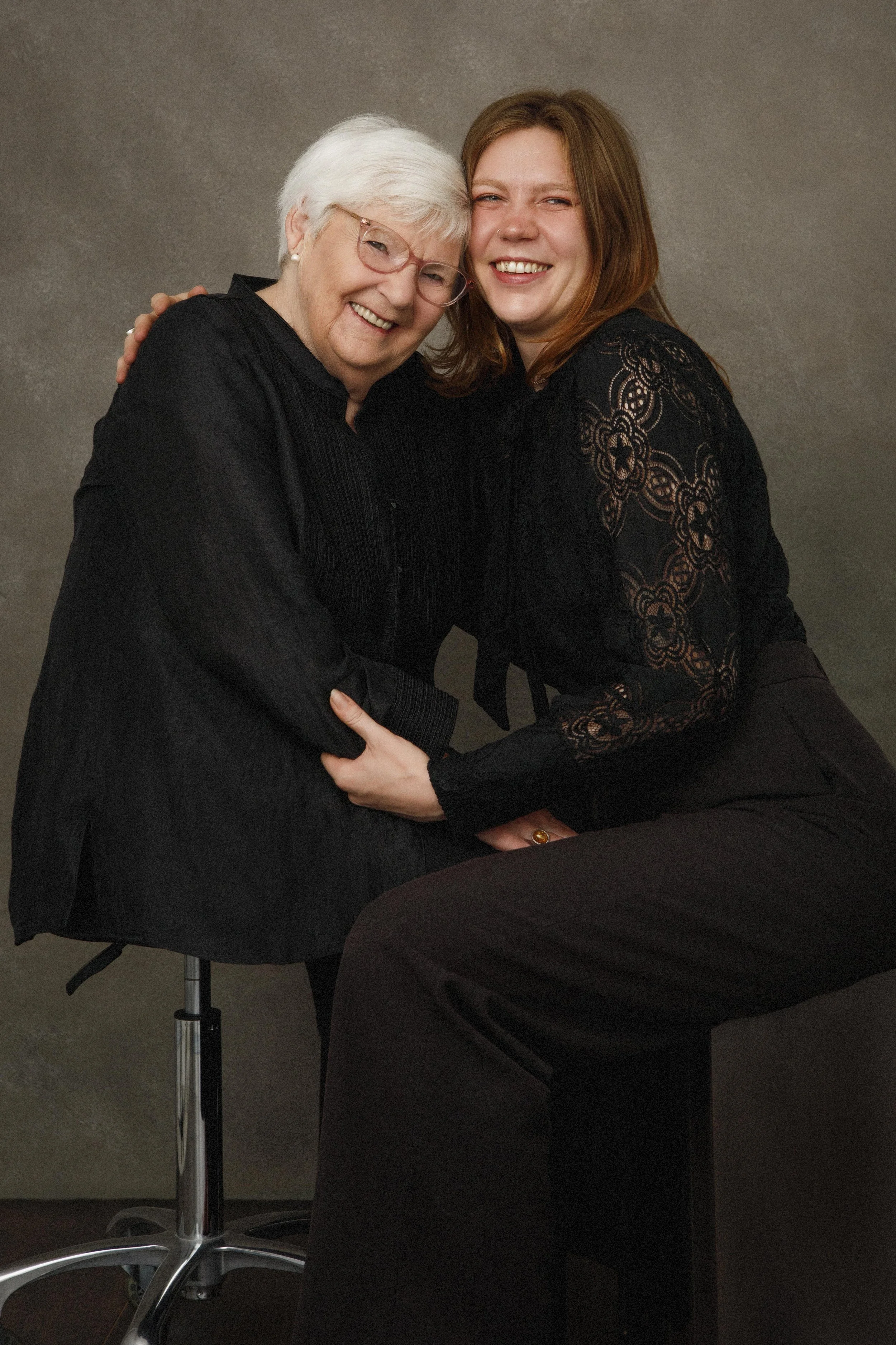 An elderly woman and a young woman hugging and smiling at the camera, with a neutral background.
