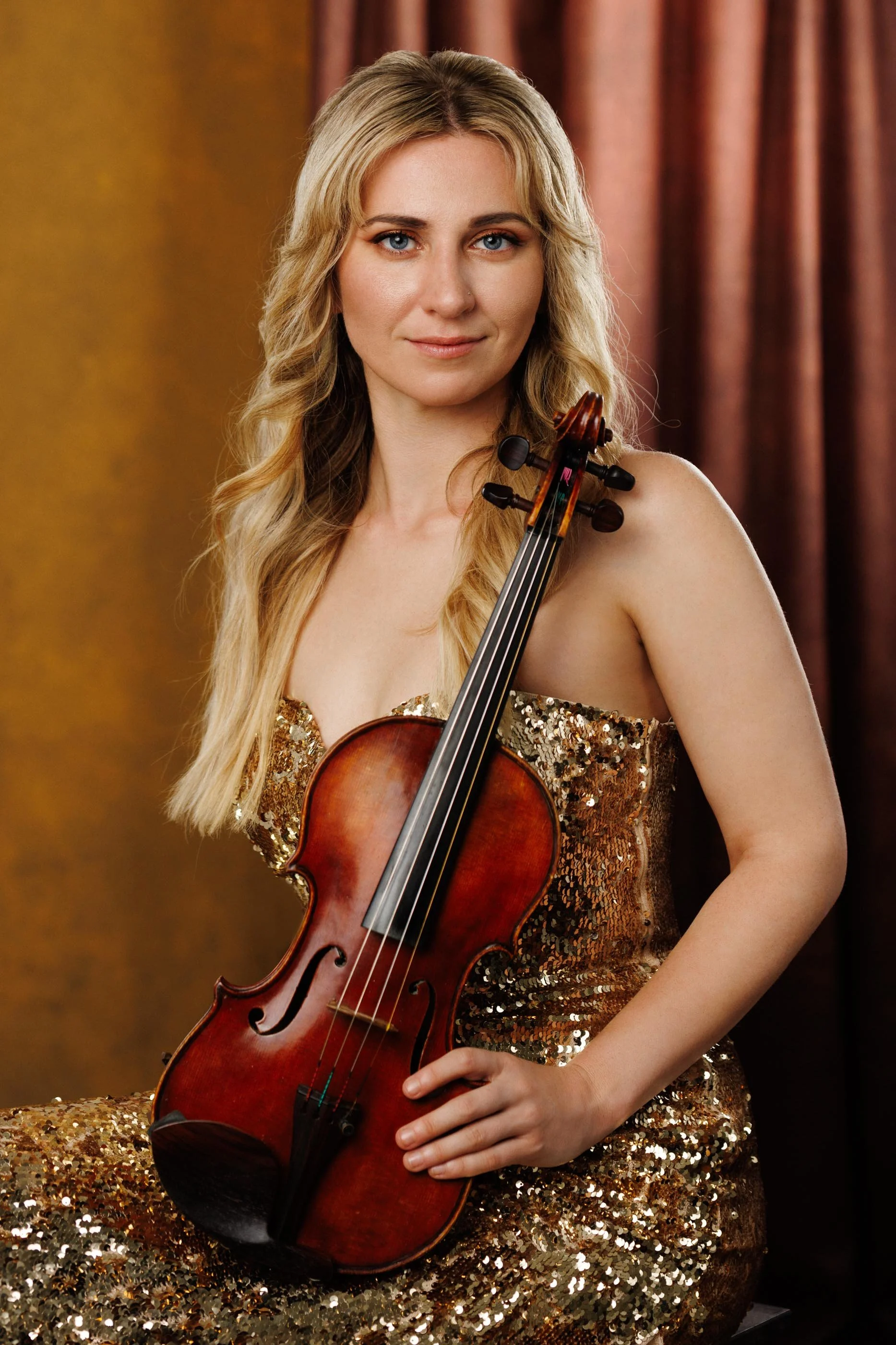 Blond hair woman wearing gold sequined dress sitting with her violin.
