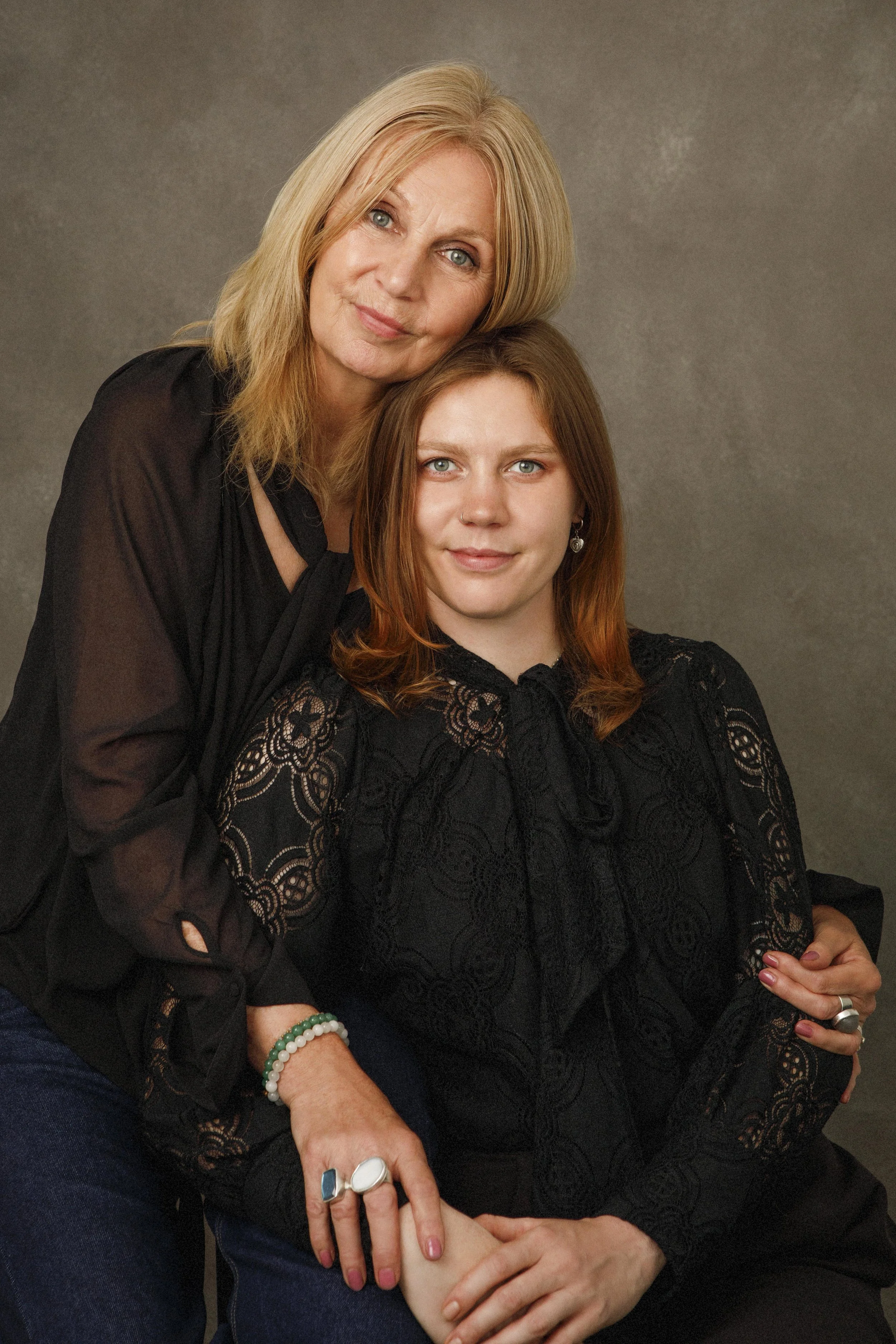 A woman with blonde hair and a woman with red hair posing together. The blonde woman is in a black sheer top and the red-haired woman is in a black lace top. The red-haired woman is sitting, and the blonde woman is leaning her head on the other's. They both have bright blue eyes and are smiling softly. The background is a plain gray surface.