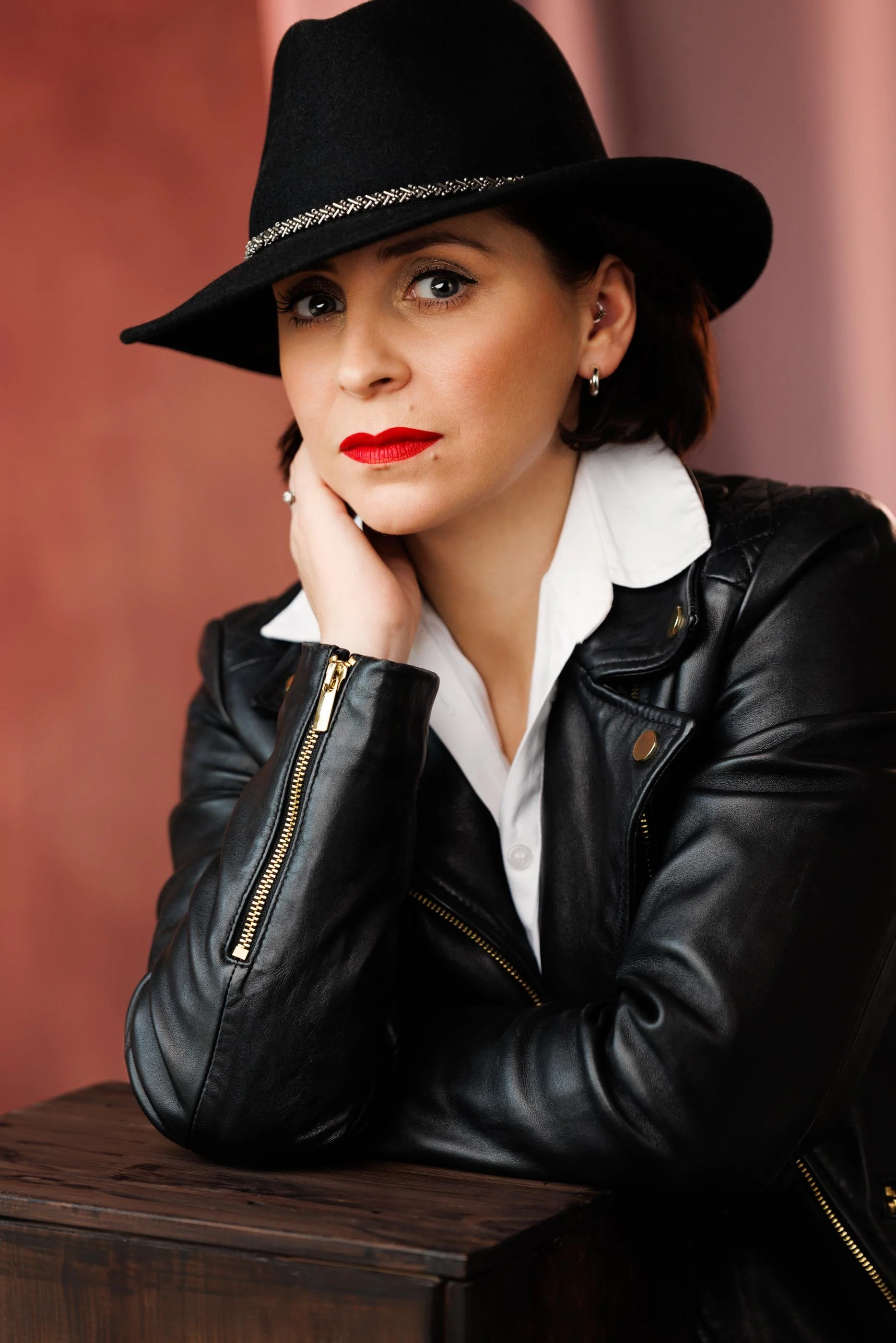 A directed studio portrait of a confident woman in a black leather jacket, white shirt, and wide-brim fedora. Red lip, direct gaze, chin resting on hand. Shot against a warm rose backdrop. The image communicates composure and self-possession — the ki
