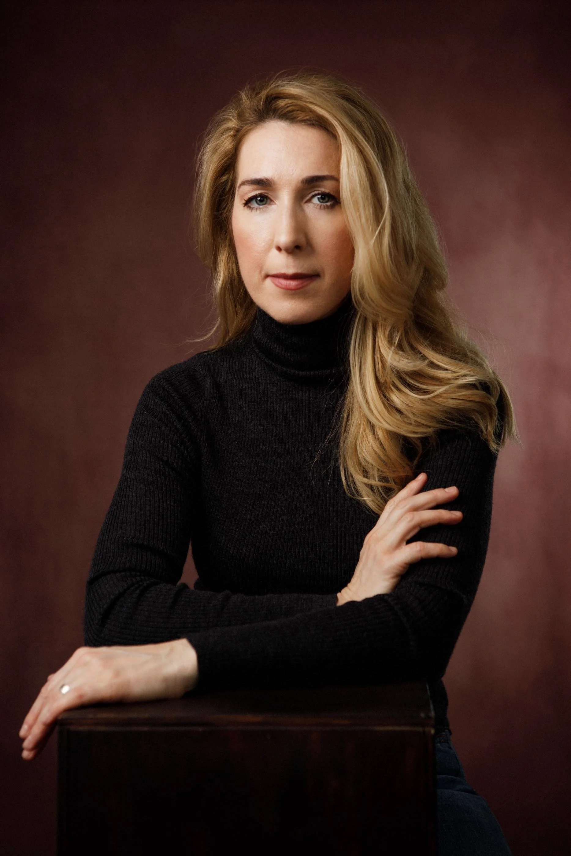 Elegant woman with blond hair and black roll neck looking into camera with dusty mauve background