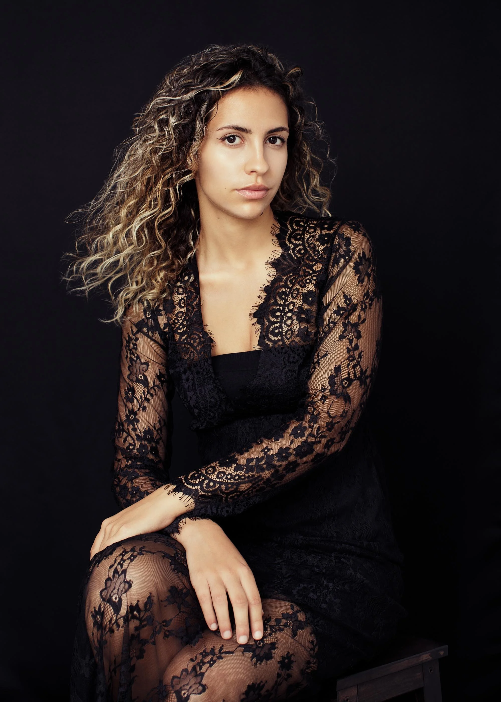 A dramatic studio portrait of a woman with natural curly hair in a full black lace dress, shot against a deep black backdrop. Low, sculpting light. Seated, hands relaxed on knees, direct unflinching gaze. The image has an editorial weight — fashion-q
