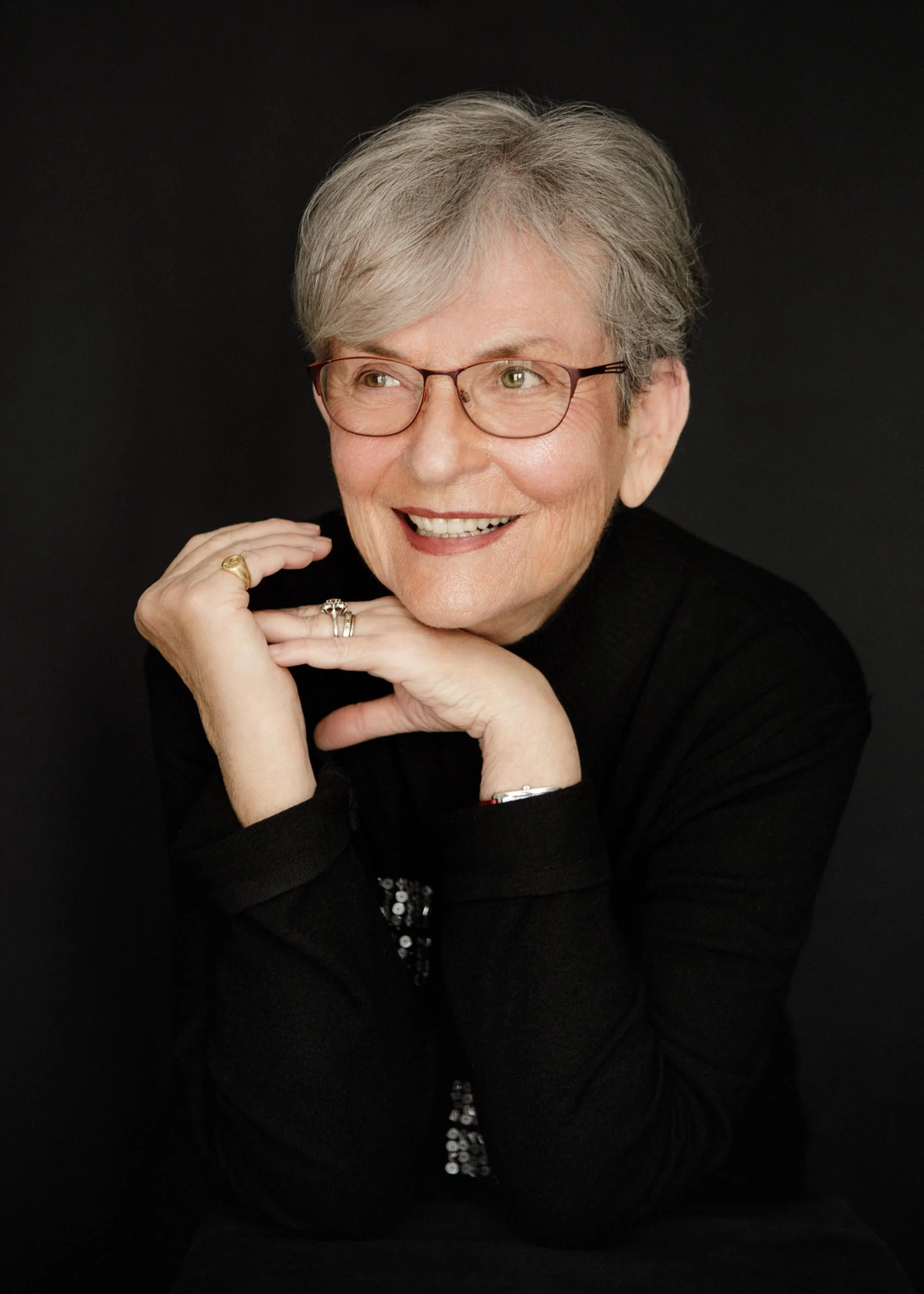 A portrait of an elderly woman with short gray hair wearing glasses, smiling, and resting her chin on her hands, against a black background.