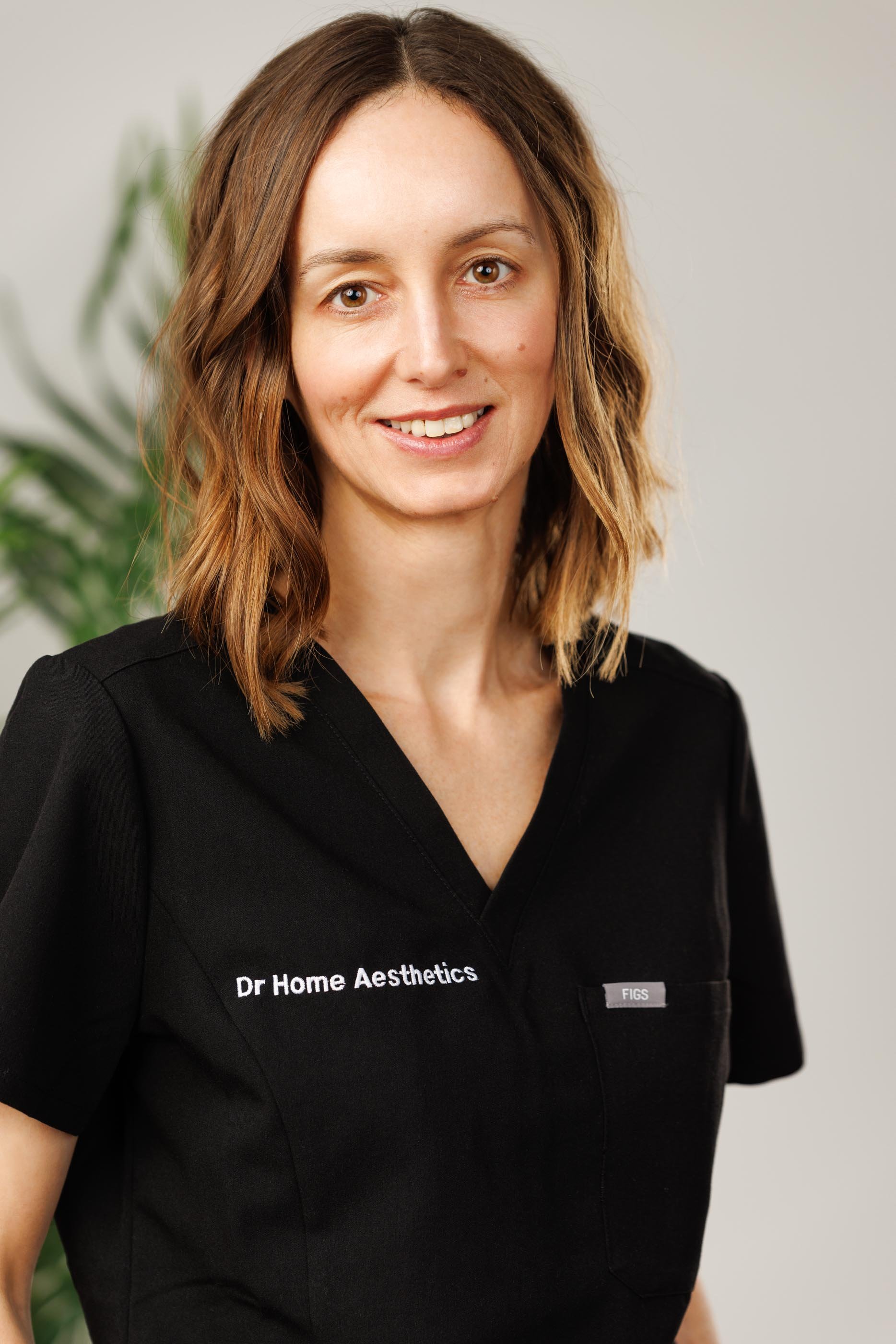 A natural light portrait of a clinic director in branded black scrubs, warm smile, relaxed and confident posture against a clean bright background with foliage. This is the second image from the same professional session — demonstrating the value of 