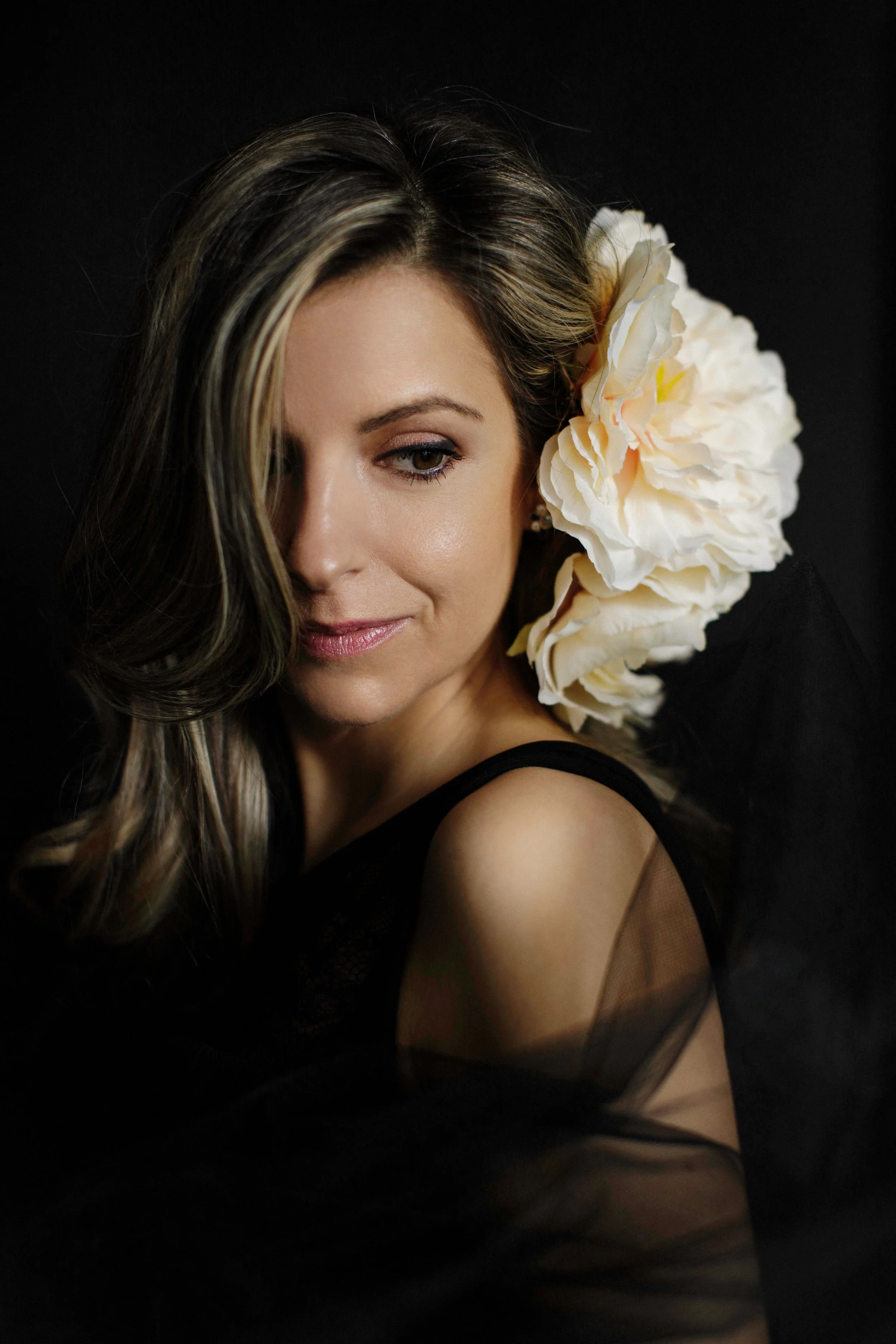 A moody, intimate studio portrait of a woman with highlighted hair, a large white peony pinned at the side, wearing a sheer black dress against a near-black backdrop. Downward gaze, soft smile. The contrast between the dark environment and the single