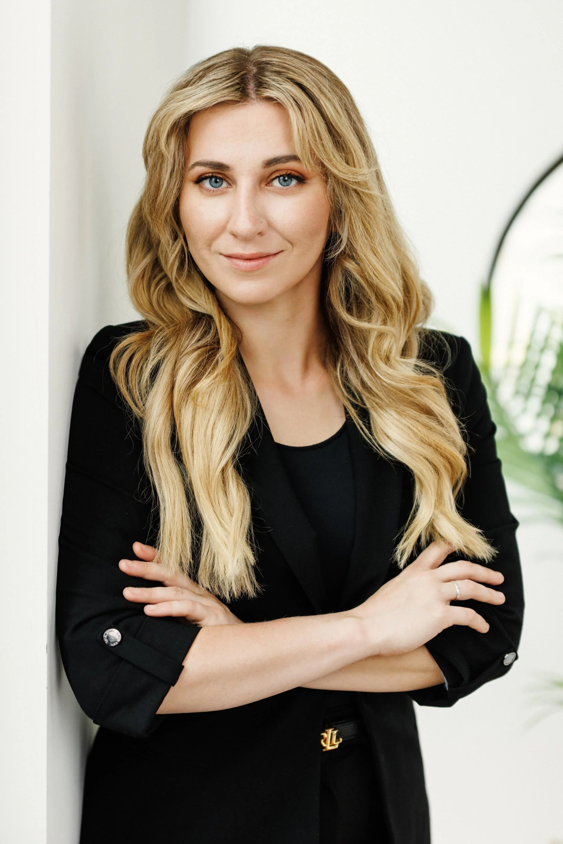 A woman with long blonde hair and blue eyes, wearing a black blazer, standing with her arms crossed, smiling and looking directly at the camera, indoors with a white wall and a green plant in the background.