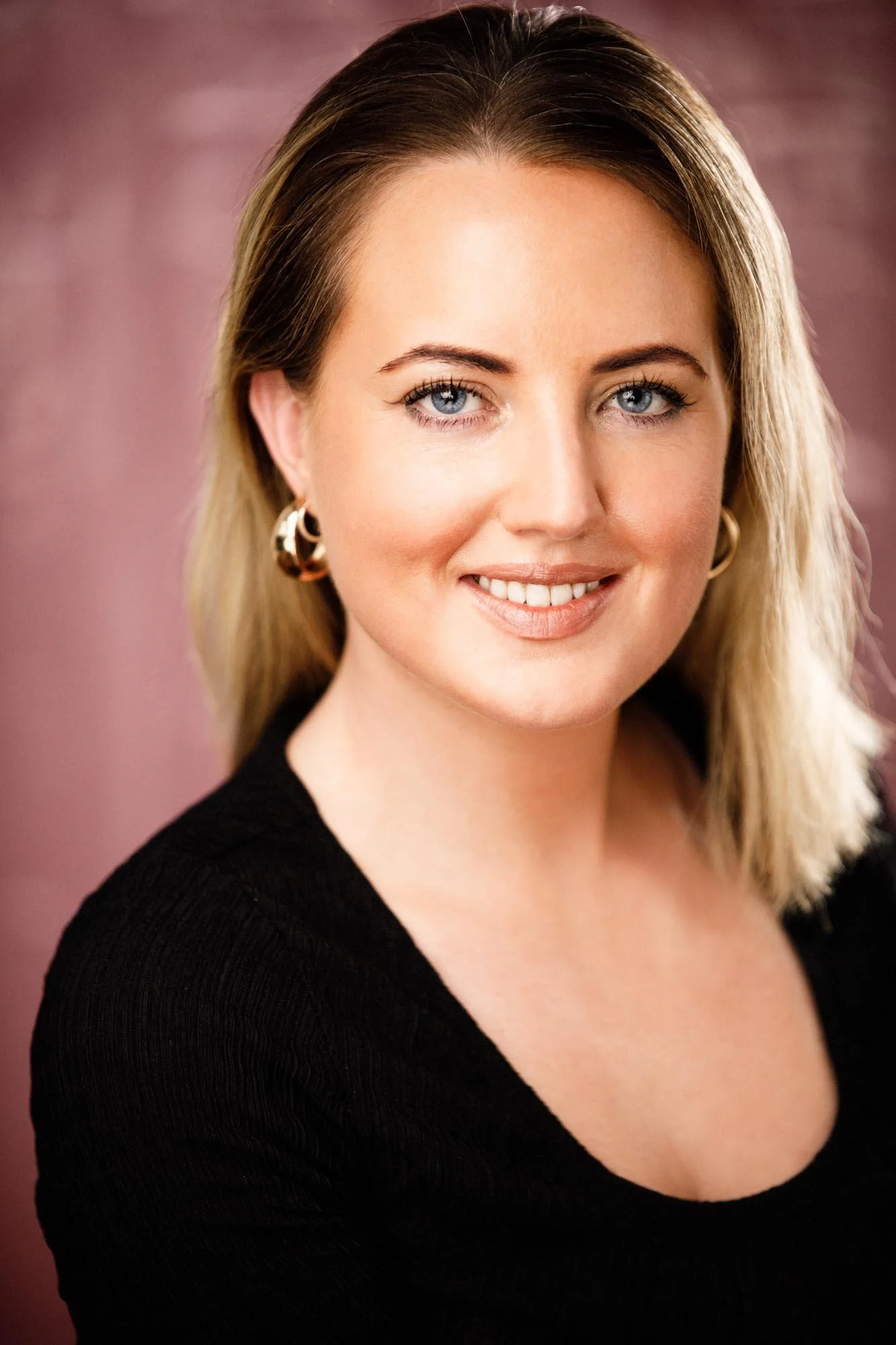 A clean, close-crop professional headshot of a blonde woman with blue eyes in a black knit top, shot against a warm rose backdrop. Gold hoop earrings, fresh makeup, genuine open smile. Bright, flattering light. This is the portrait that performs cons