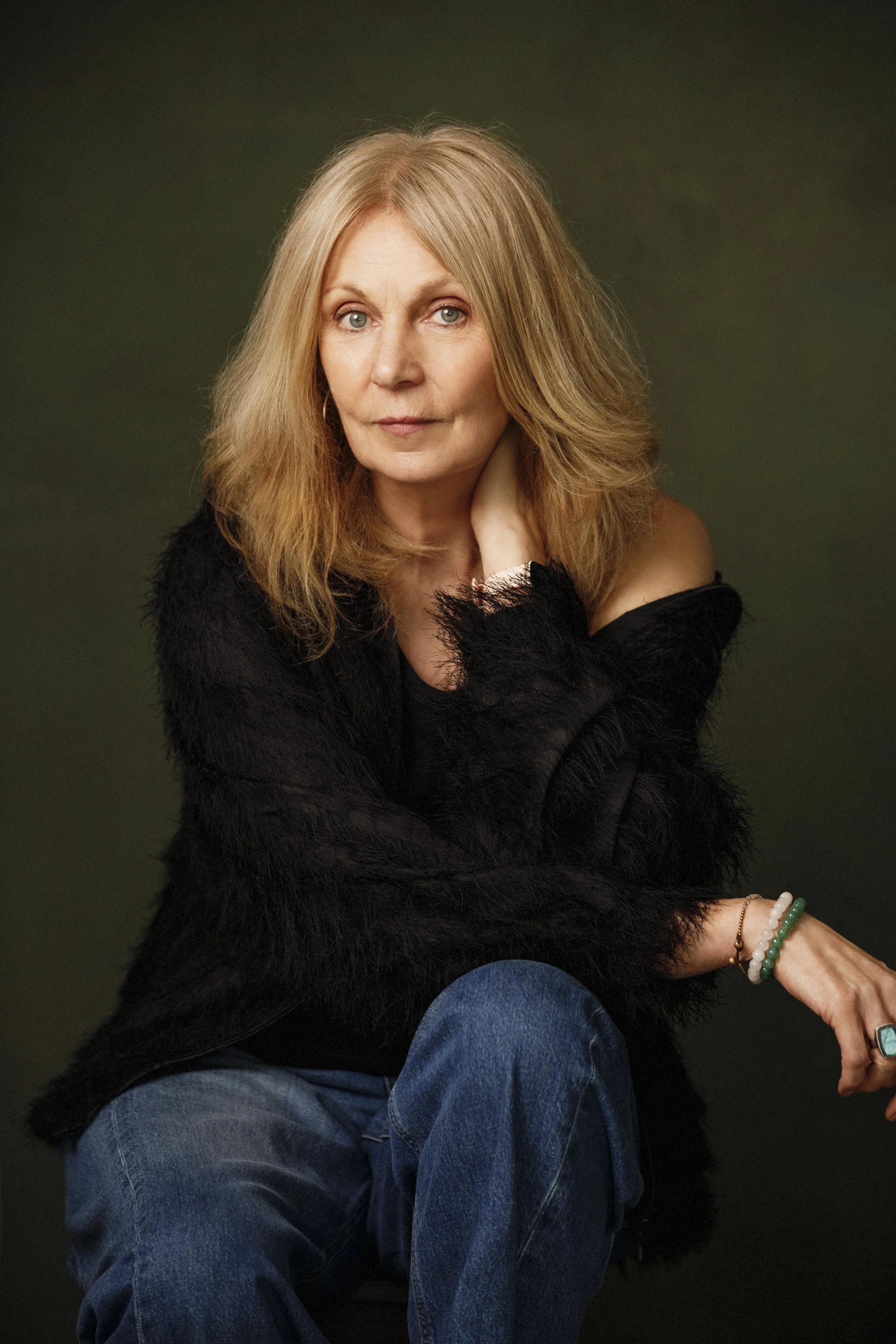 A portrait of a mature woman with long, wavy blonde hair, wearing a black fuzzy sweater, jeans, and bracelets, sitting with her hand near her neck against a dark background.