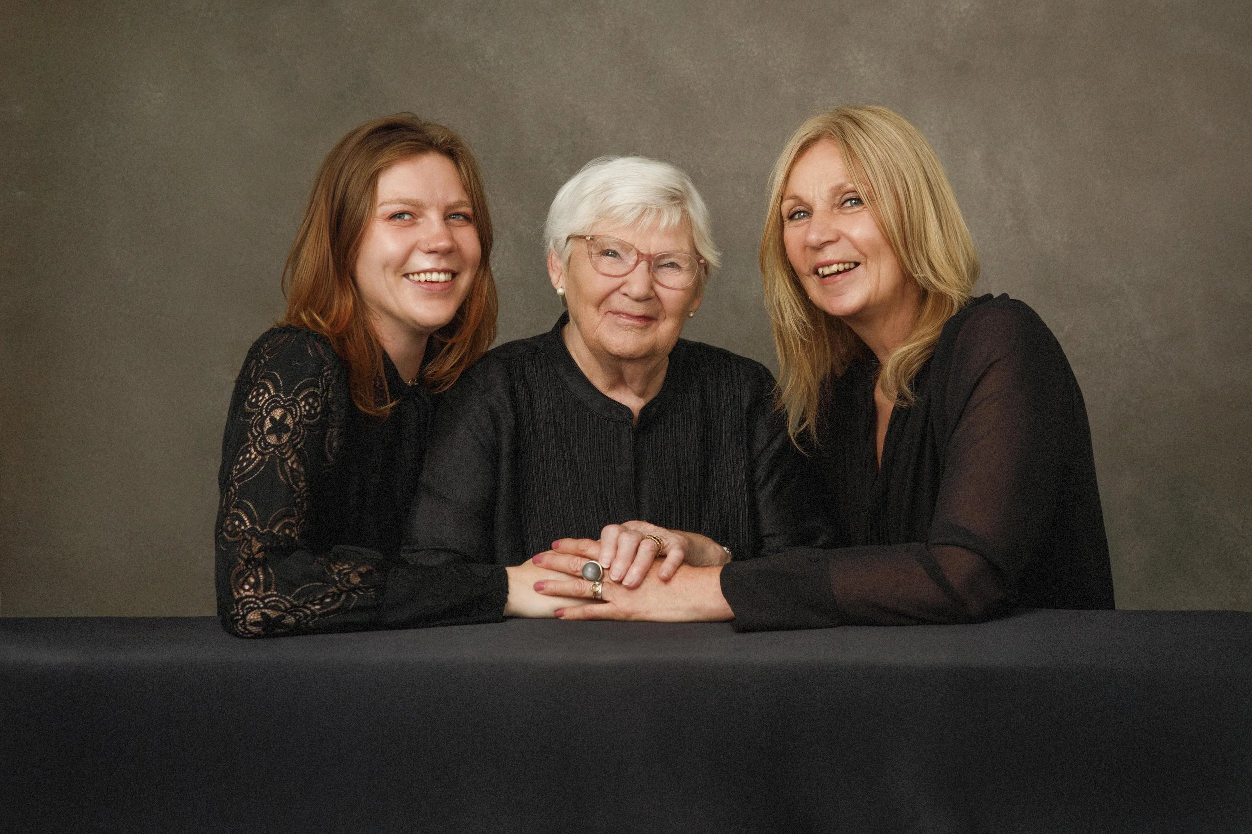Three women sitting together, holding hands on a table, smiling, against a plain grey background.