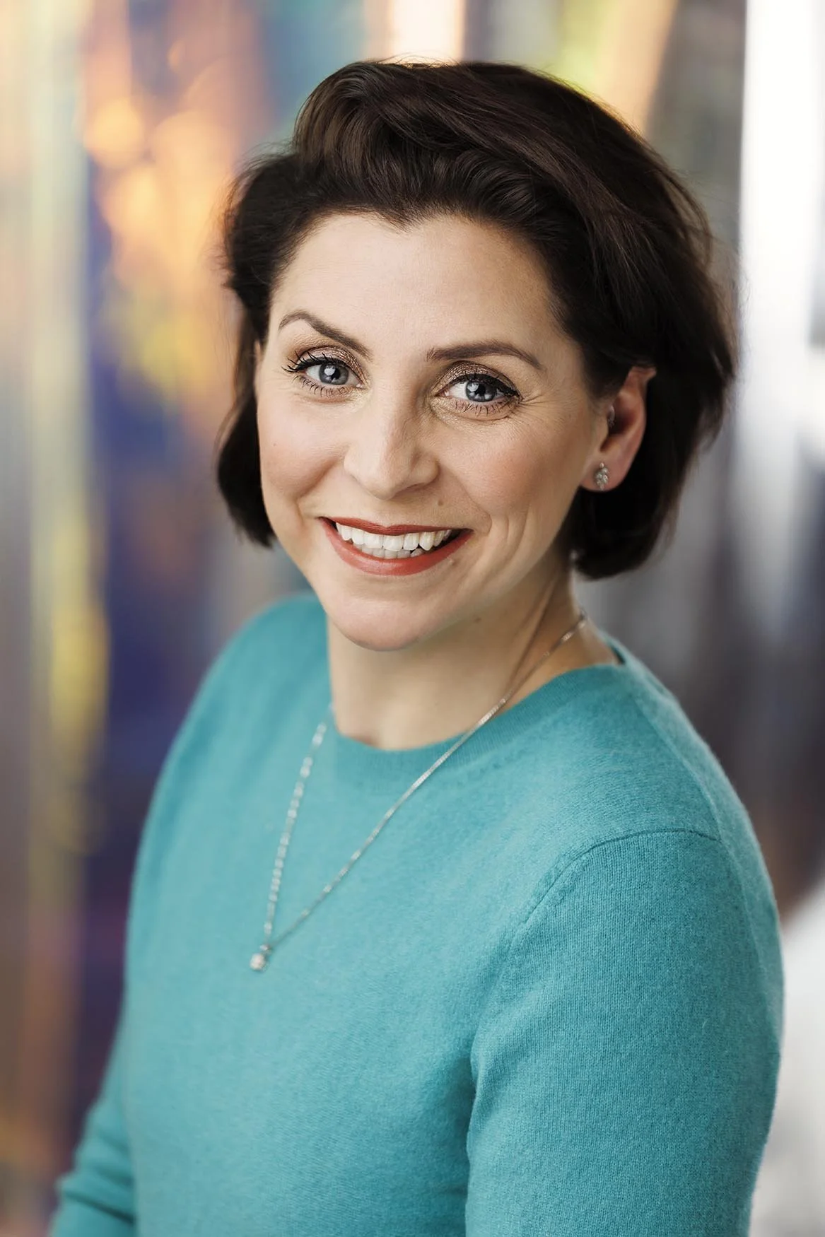 A bright, close-crop portrait of a dark-haired woman with blue eyes in a teal sweater, shot against a softly blurred colourful backdrop. Warm, genuine smile, delicate necklace, understated styling. This is the portrait for the professional woman whos