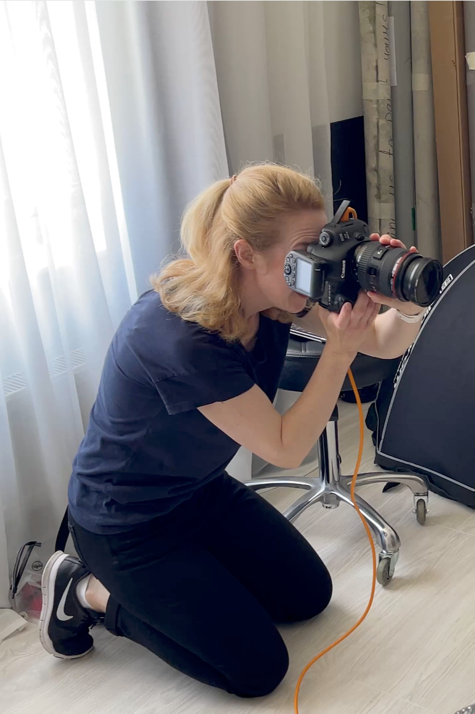 A woman kneeling on the floor taking a photograph with a professional camera in an indoor setting with white curtains and a wooden cabinet.