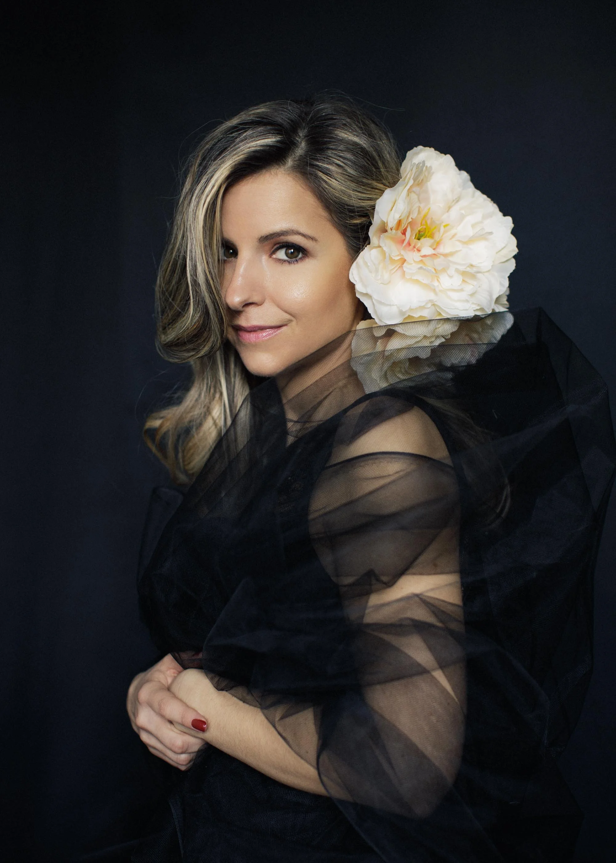 A woman with a white flower in her hair and wrapped in a black tulle, looking sideways to the camera.