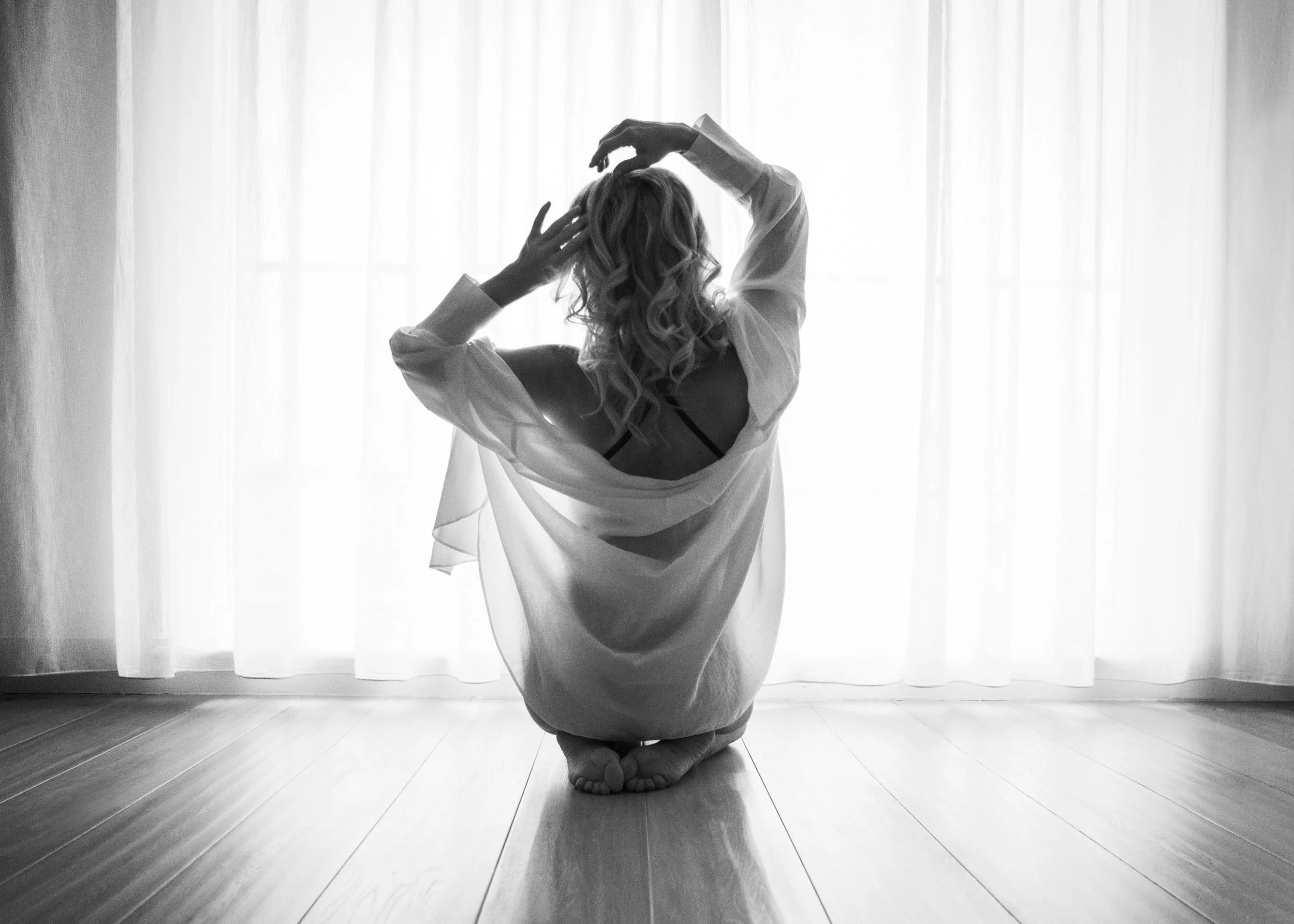 A black and white fine art boudoir portrait of a woman seated on a wooden floor in a sheer robe, shot from behind against floor-to-ceiling backlit curtains. Arms raised, curly hair loose, silhouette defined by natural window light. Anonymous, intimat
