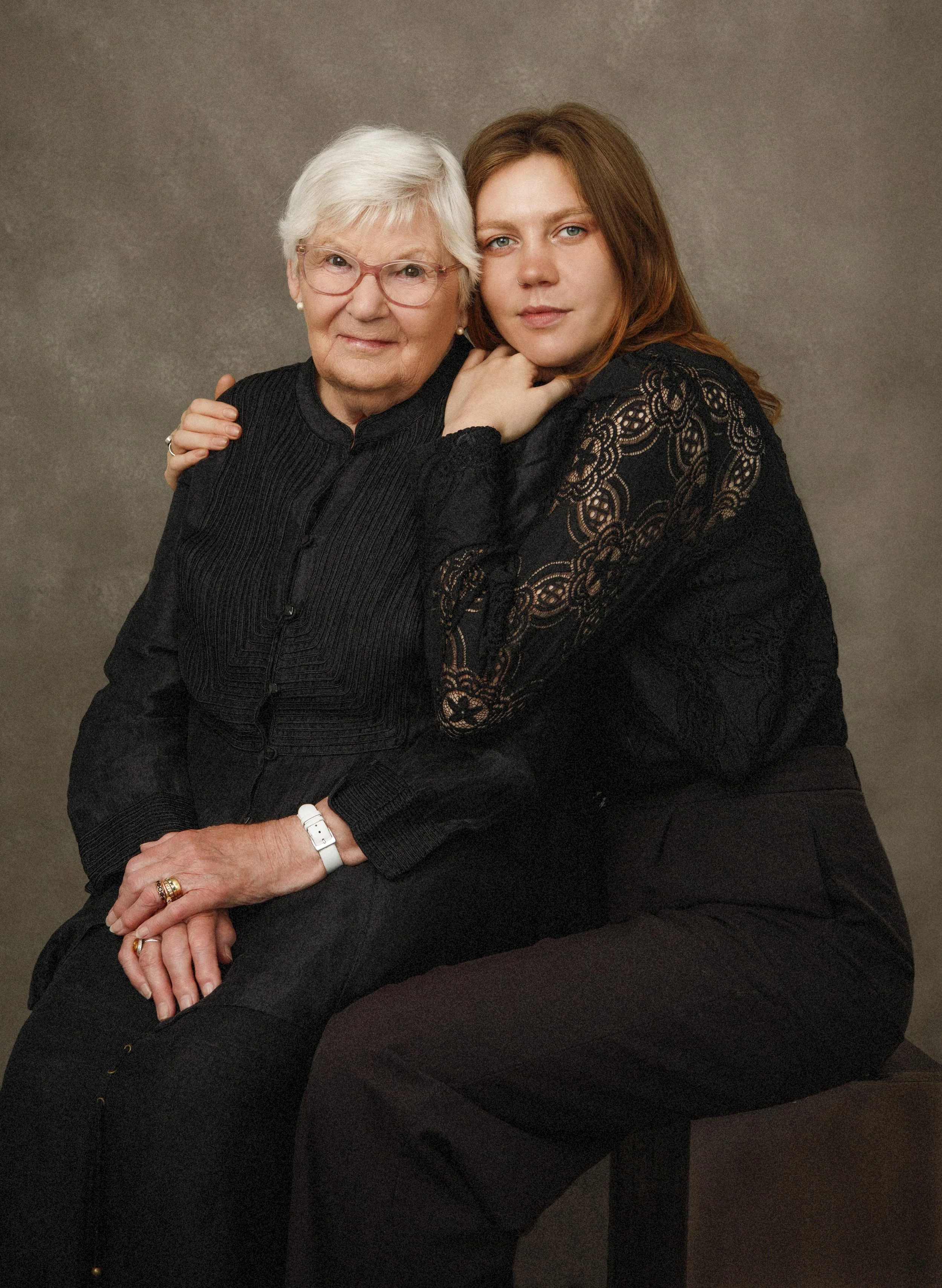 A portrait of an elderly woman with white hair and glasses, and a young woman with brown hair, both wearing black clothing, sitting close together against a plain brown background.