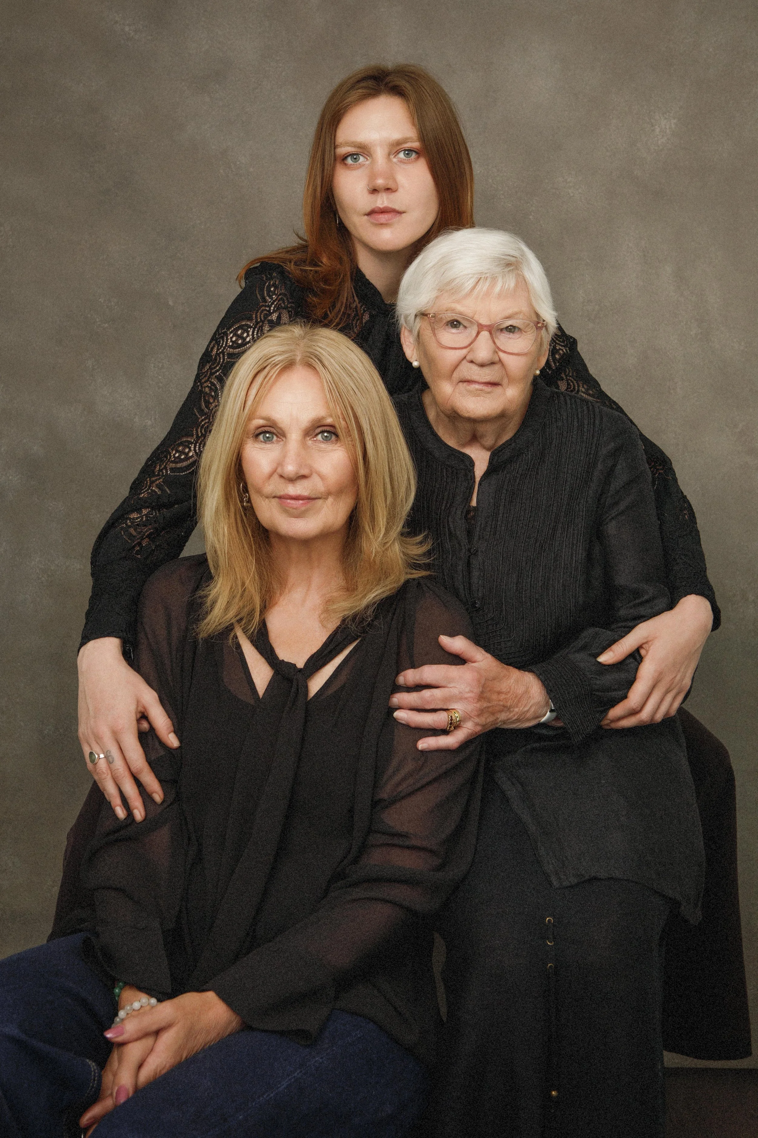 Three women sitting together, with the woman in the middle having white hair and glasses, the woman on the left having blonde hair, and the woman behind with auburn hair. They are all looking at the camera against a plain gray background.