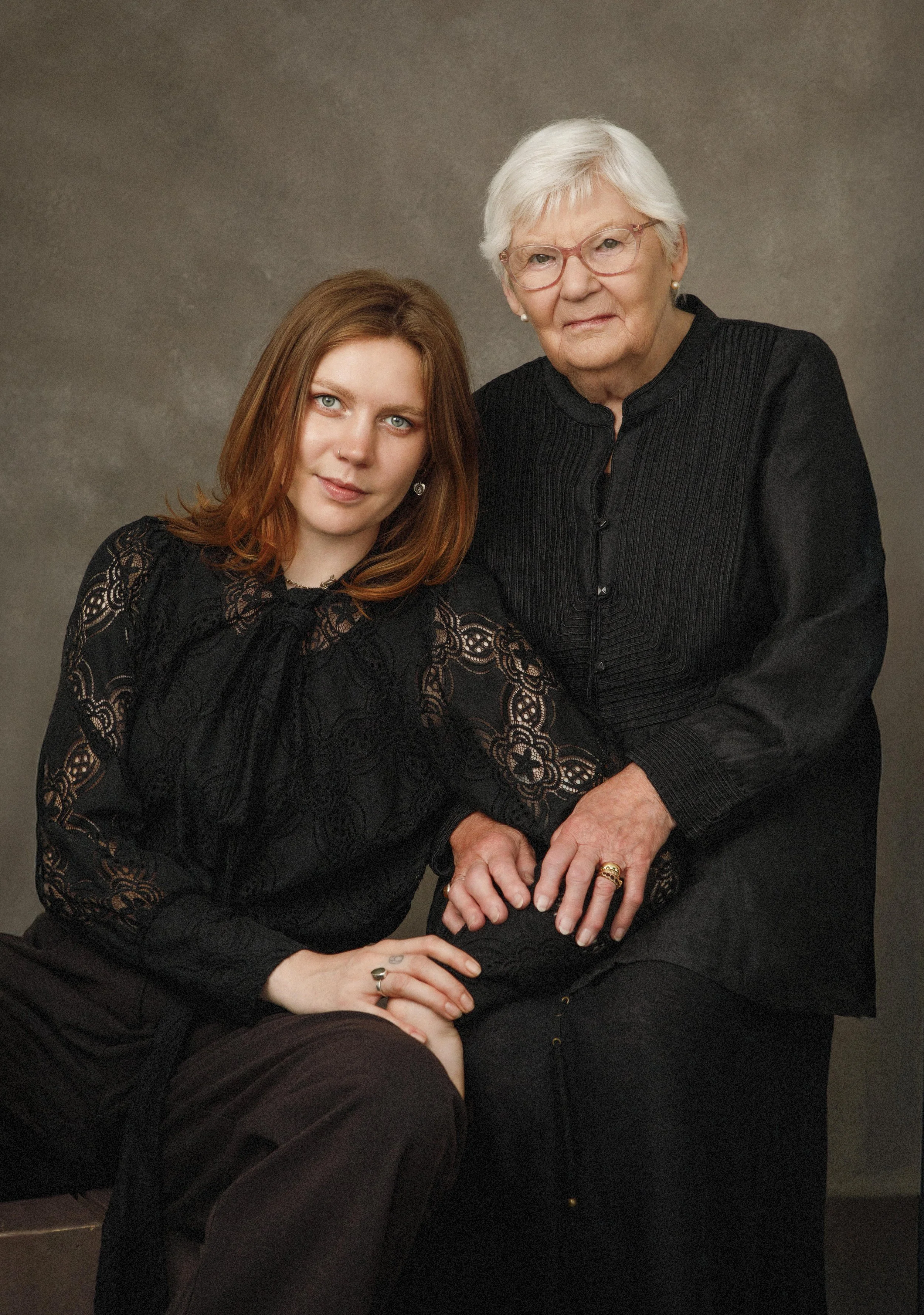 A young woman with red hair and blue eyes sitting next to an elderly woman with white hair and glasses. They are dressed in dark clothing and sitting close together against a plain background.