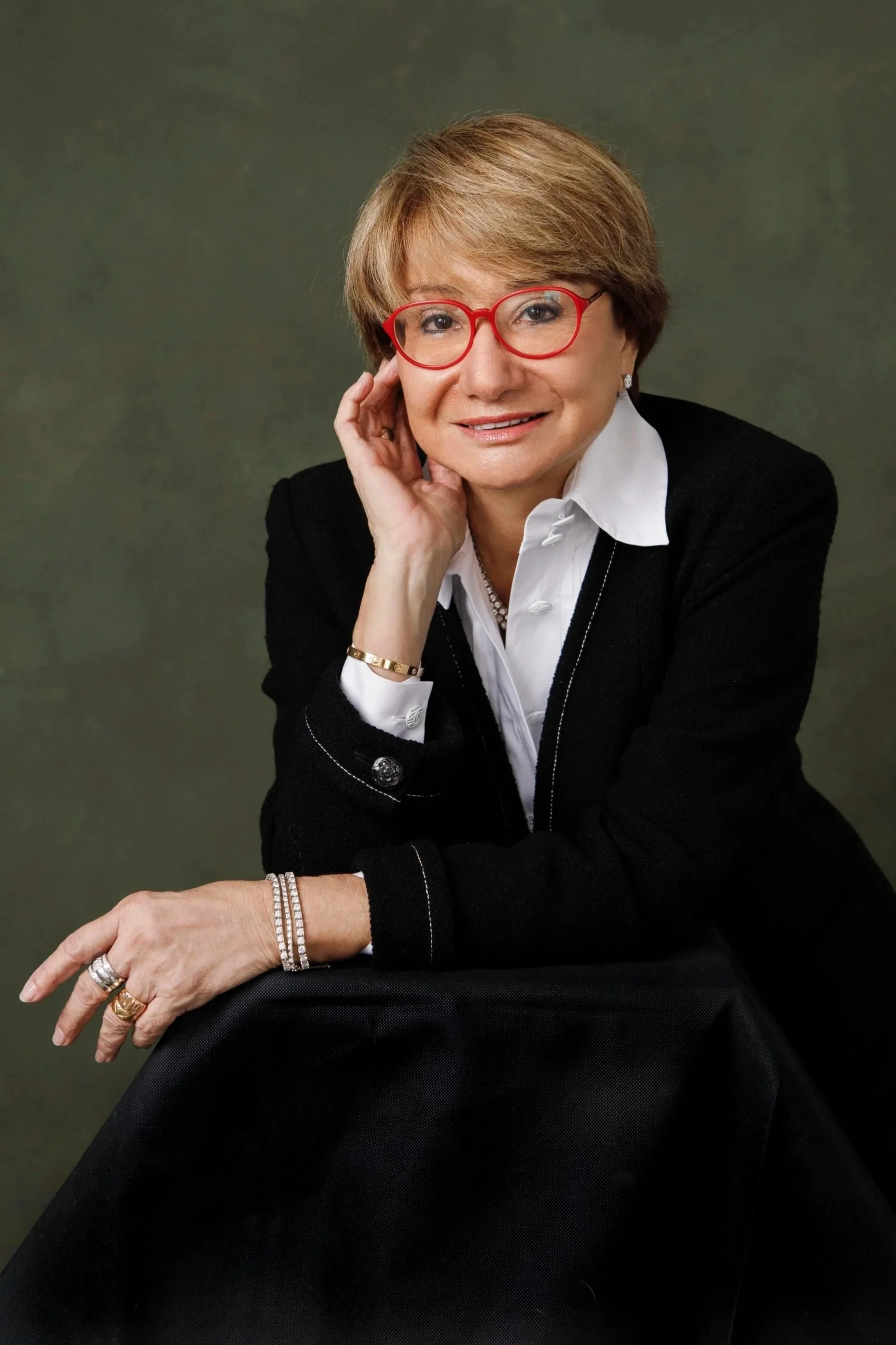 A smiling middle-aged woman with short brown hair wearing red glasses, a white collared shirt, and a black jacket, posing with her hand resting on her face and her left arm on her knee, against a dark green background.