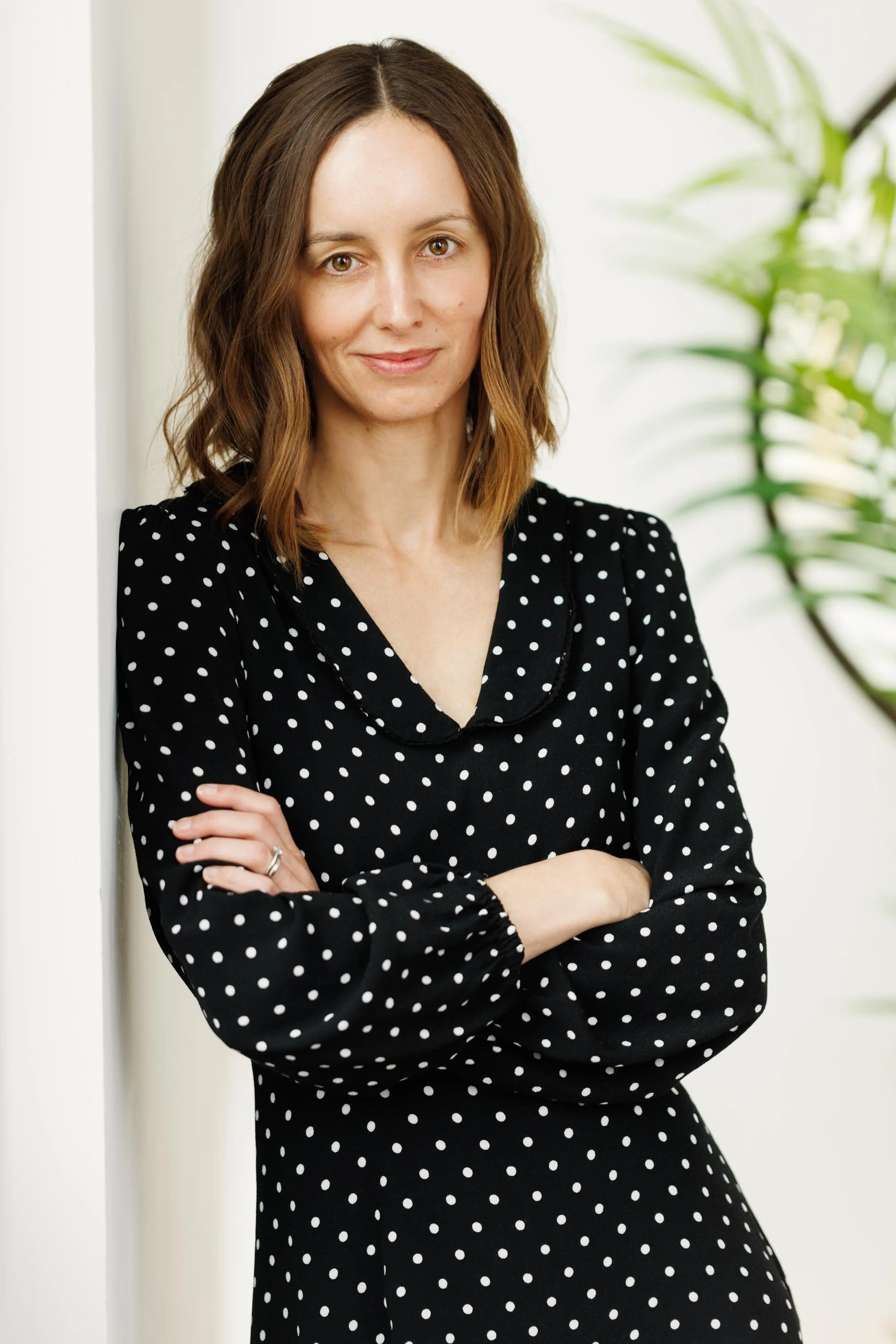 A clean, natural light portrait of a professional woman in a black polka dot wrap dress, arms crossed, leaning slightly with a calm, direct expression. Bright background with foliage, relaxed environment. This image sits at the intersection of person