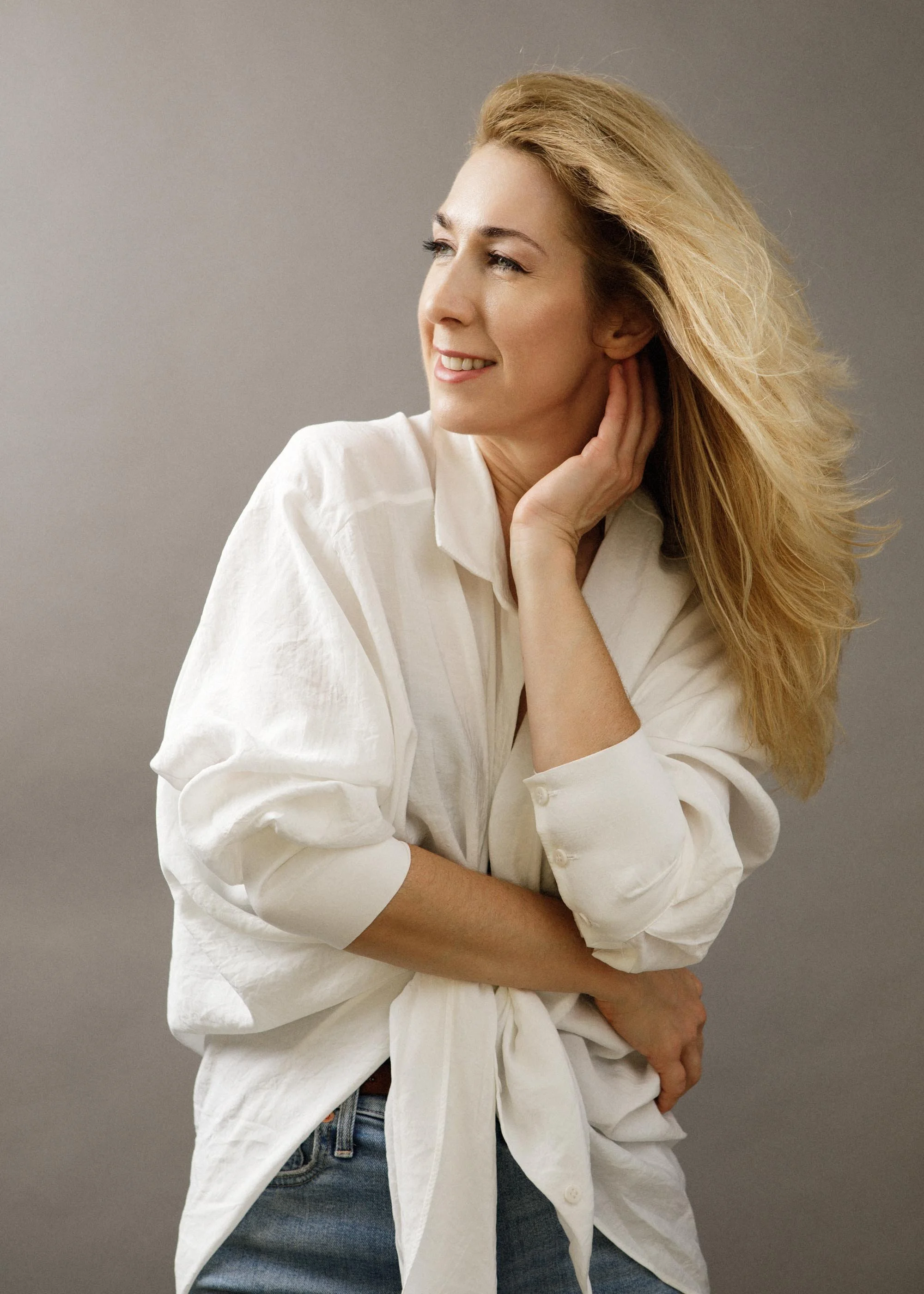 A woman with long, wavy blonde hair gazing to her left, smiling softly, wearing a white button-up shirt and blue jeans, against a neutral gray background.