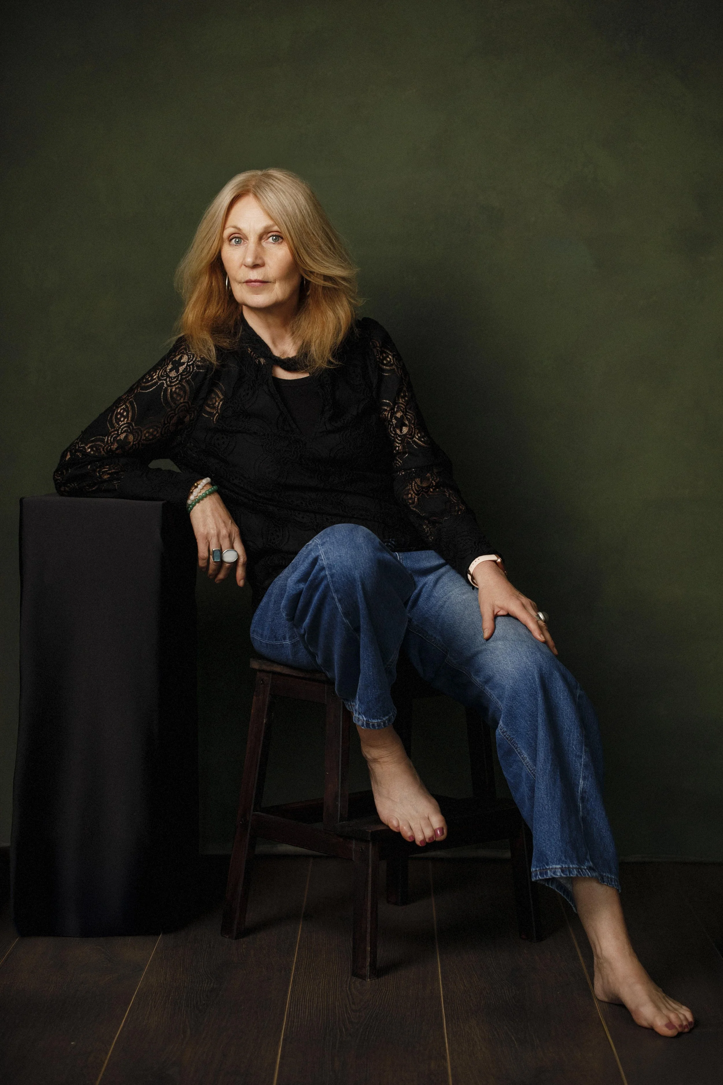 A middle-aged woman with long, blonde hair and blue eyes, wearing a black lace top and blue jeans, sitting on a wooden stool with one leg crossed over the other, barefoot, against a dark green background.