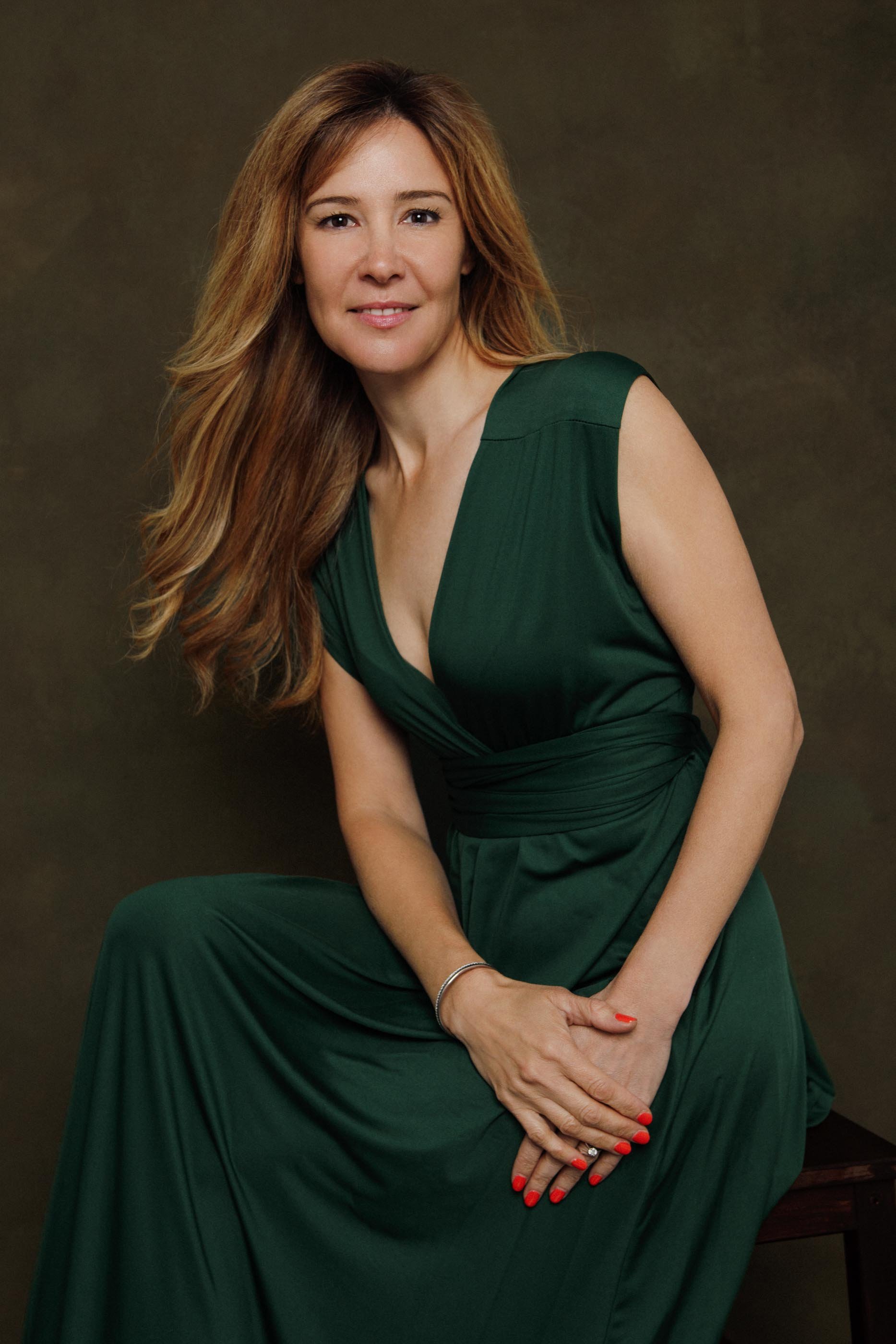 
A warm studio portrait of a woman with long auburn hair, seated in a floor-length emerald green wrap dress against a deep olive backdrop. Hands folded, relaxed posture, natural smile. The colour palette is deliberate — rich, confident tones that com