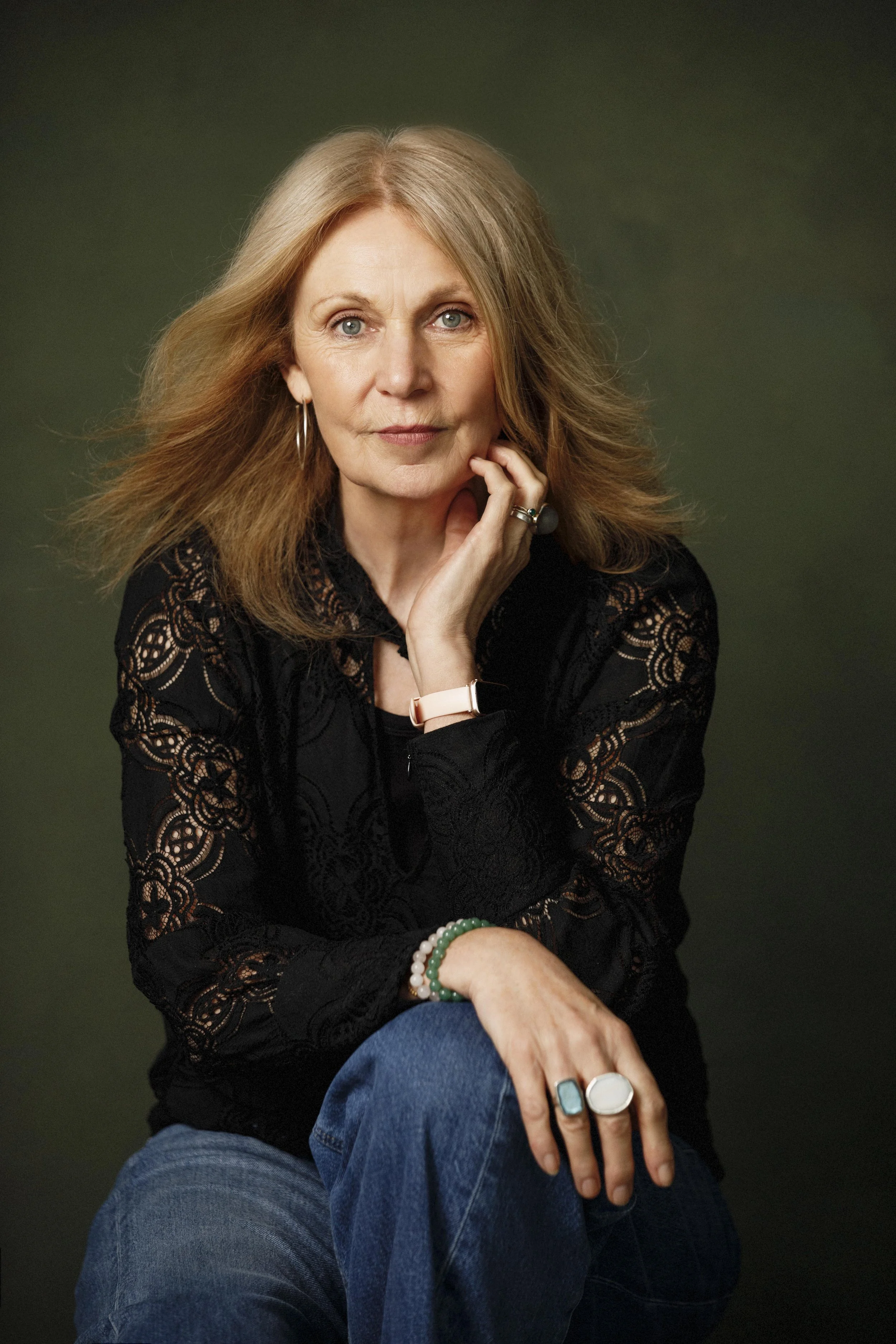 Portrait of a woman with blonde hair, wearing a black lace top, blue jeans, and jewelry, sitting against a dark green background.