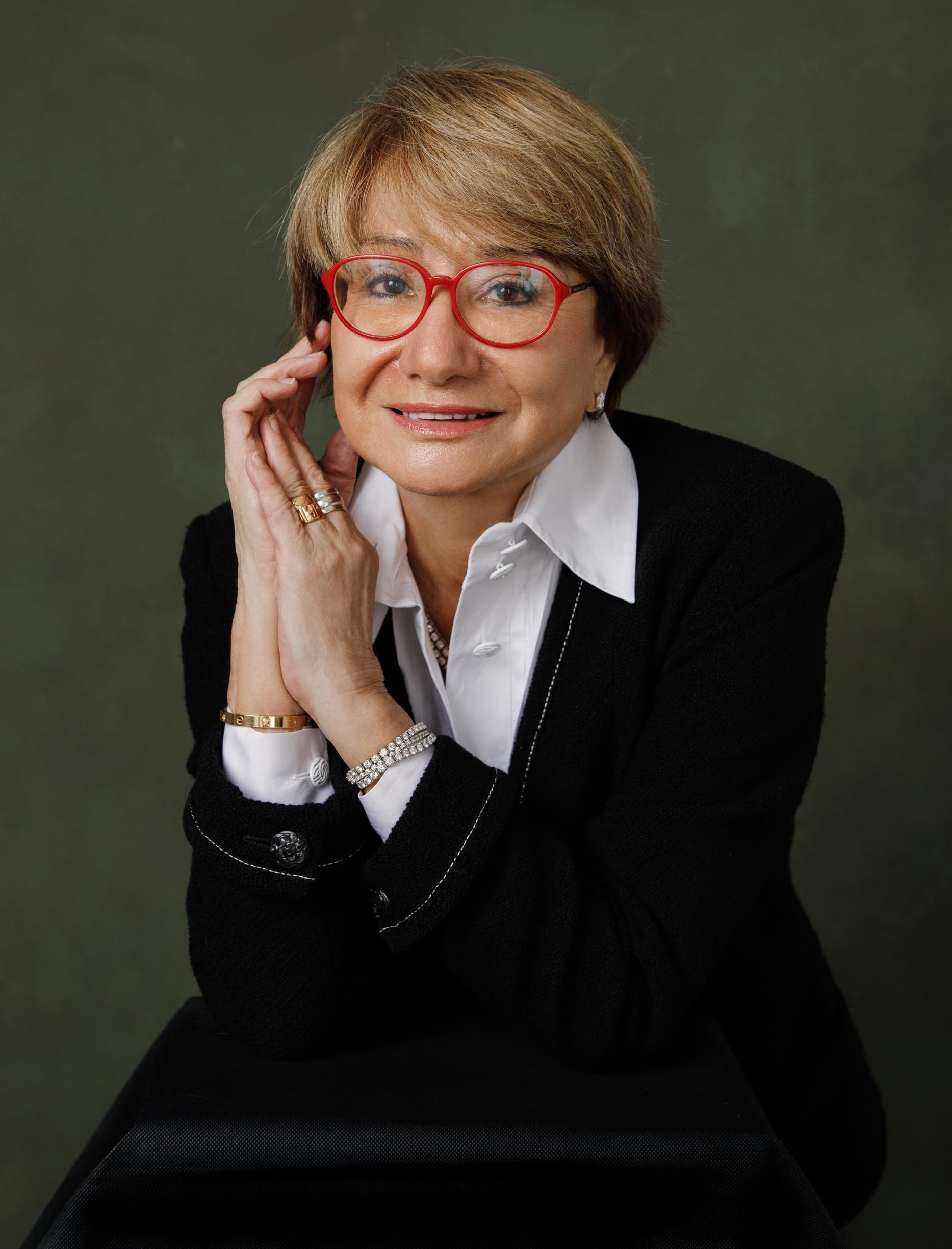 A polished studio portrait of a senior professional woman in a black blazer, white shirt, and signature red-frame glasses. Warm green backdrop, hands folded at chin, composed and assured expression. Gold jewellery, precise styling. This image represe