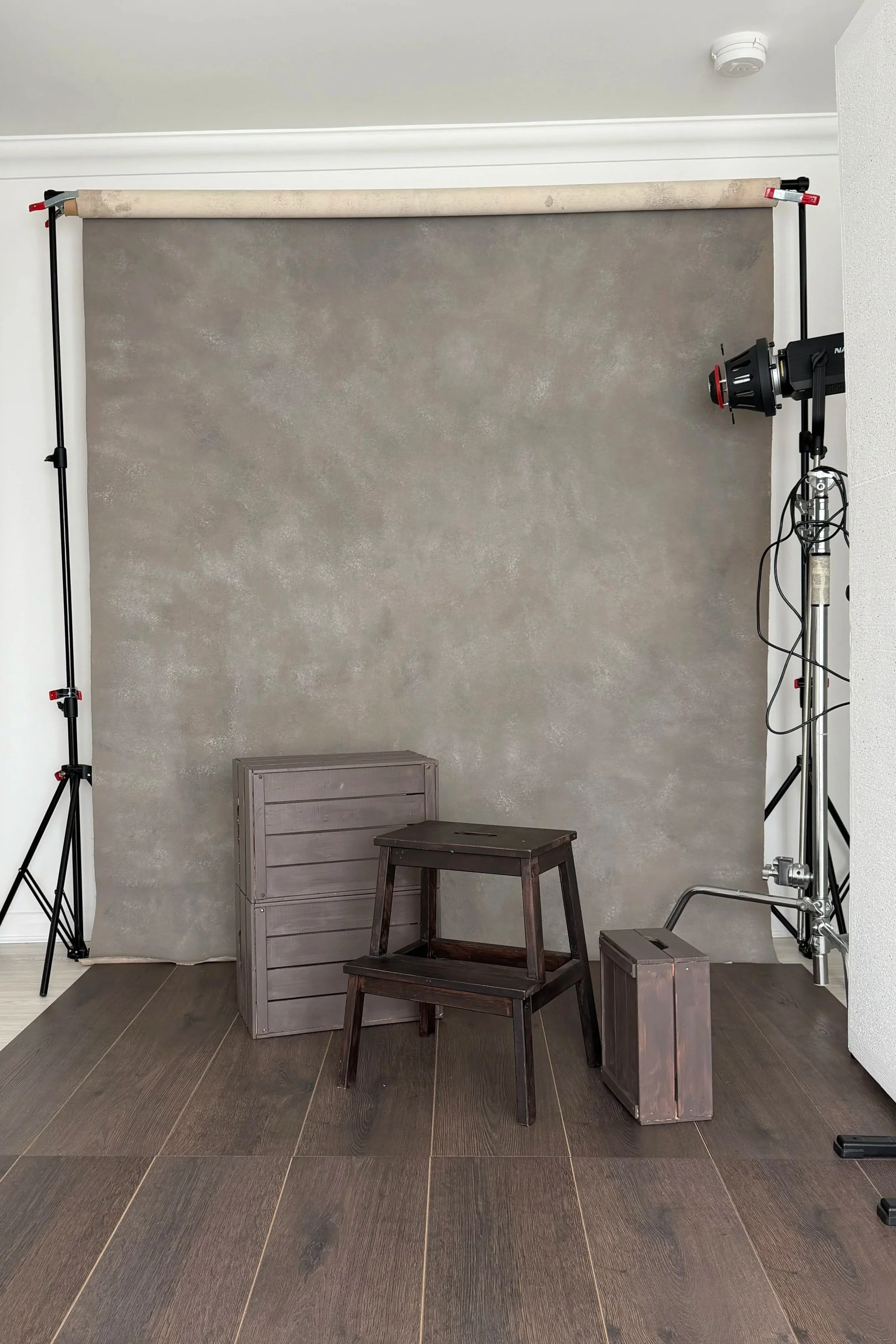 A photography studio setup with a gray textured backdrop, a wooden crate, a small wooden stool, and a wooden box, with lighting equipment on either side.