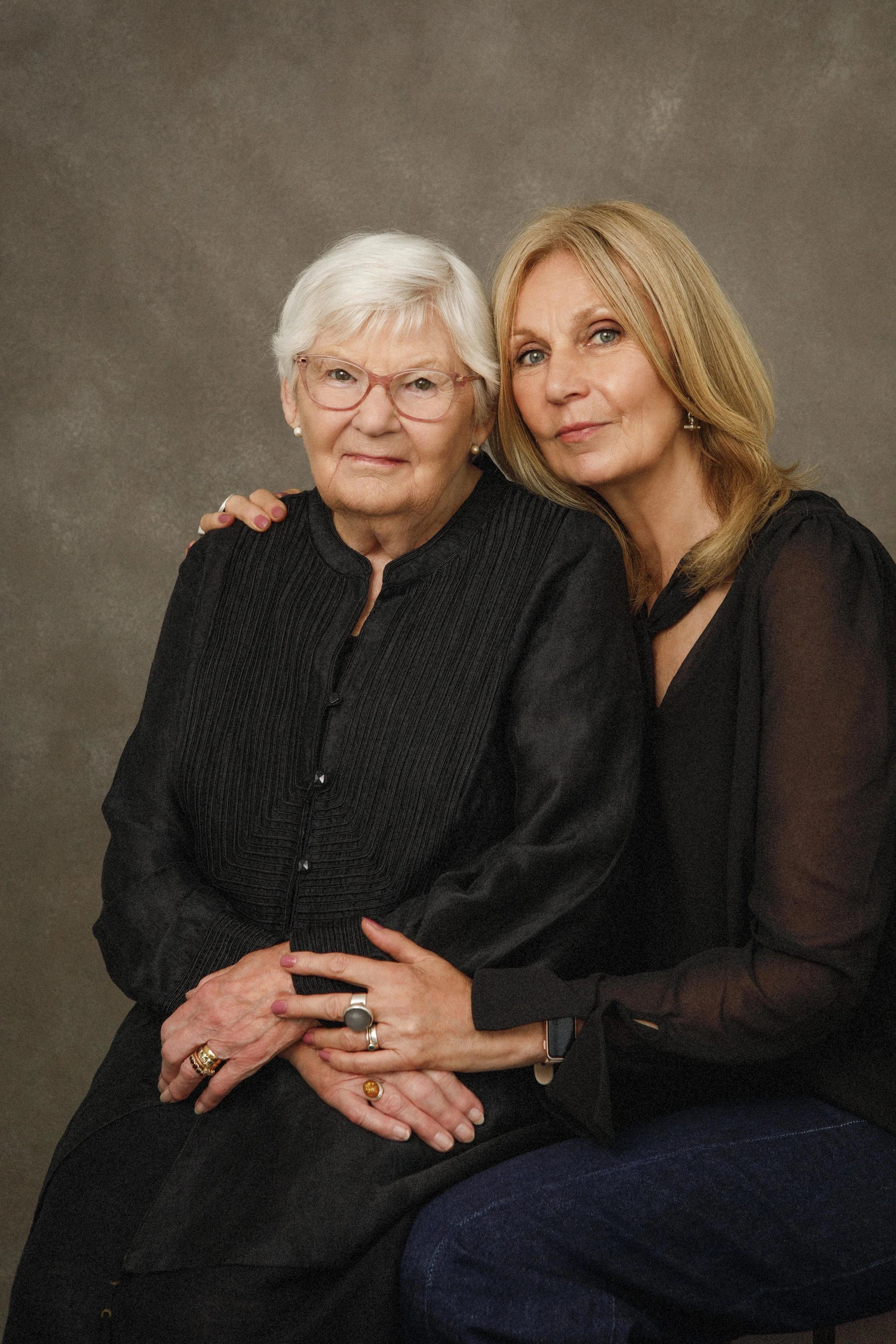 A portrait of an elderly woman with short white hair and glasses, sitting next to a middle-aged woman with long blonde hair, both wearing black clothing, against a brown background.