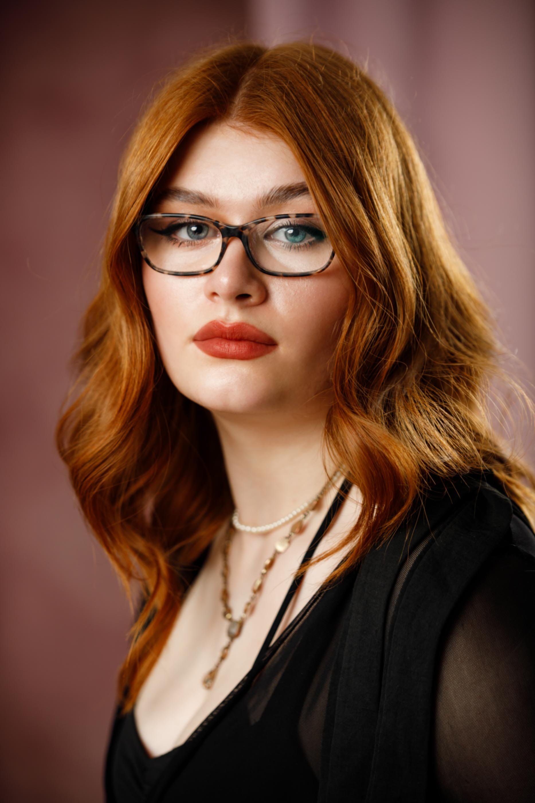 A woman with long red hair and glasses, wearing a black top and layered necklaces, looking directly at the camera against a blurred pink background.